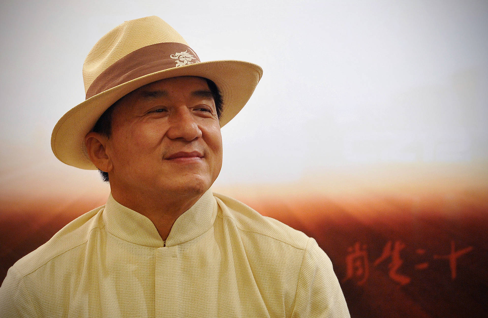 成龍戴傳統編織帽 壁紙 — 一位戴著米色帽子、穿著相配襯衫的男子，在柔和日落背景下，伴隨著中文字，溫暖地微笑。
