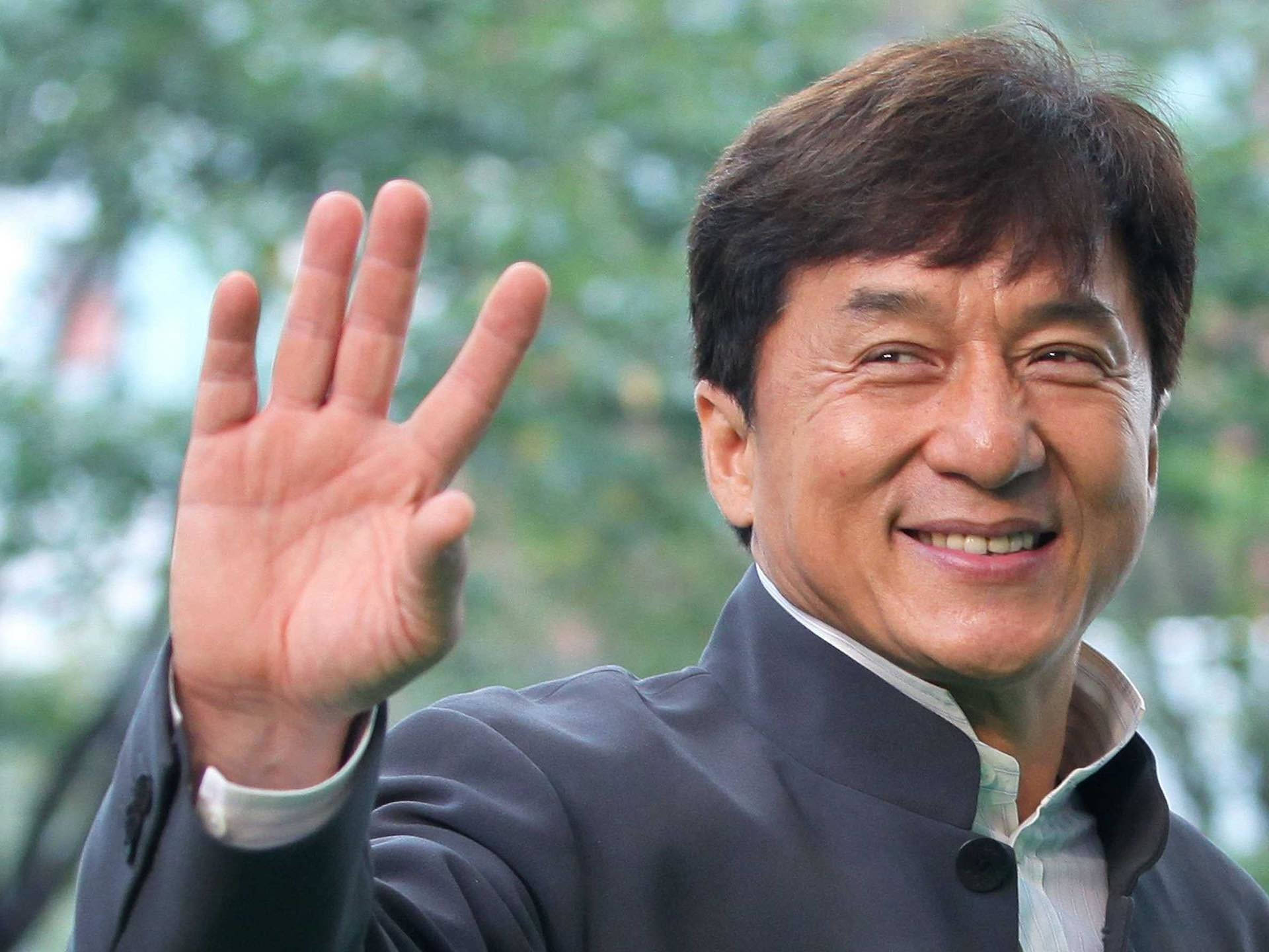成龍揮手致意 壁紙 — 一個開心的男人穿著深色外套，在柔和的綠色樹葉背景下，帶著燦爛的笑容熱情地揮手。