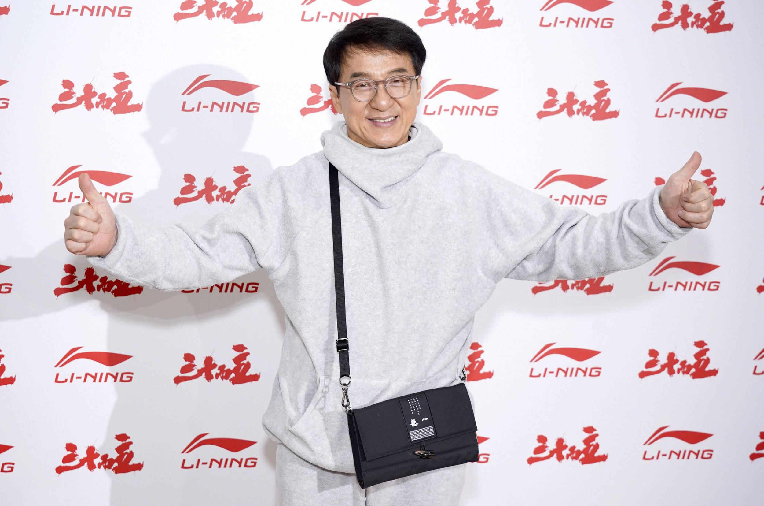 成龍身穿白色連帽衫 壁紙 — 成龍在品牌背景前穿著灰色連帽衫和黑色斜孭袋，咧嘴大笑並豎起兩個大拇指。