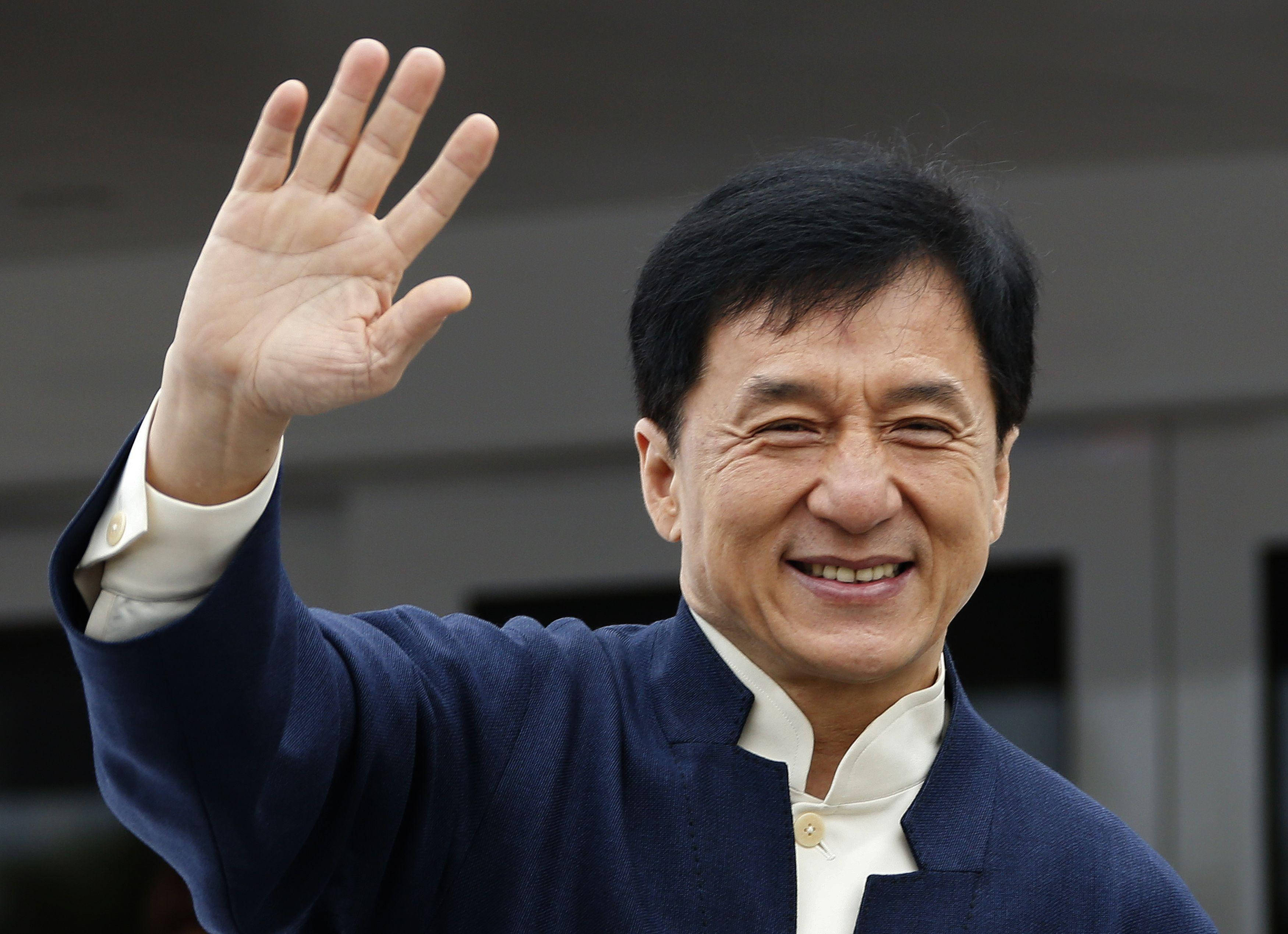 成龍開心揮手 壁紙 — 成龍身穿海軍藍外套，在模糊的現代建築背景下，帶著溫暖的笑容愉快地揮手。