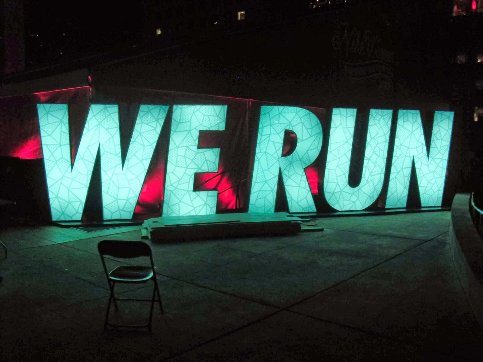 我們奔跑 壁紙 — 巨大的發光青綠色字母拼出WE RUN，在夜間以紅色背光照明，前景有一張孤獨的椅子。