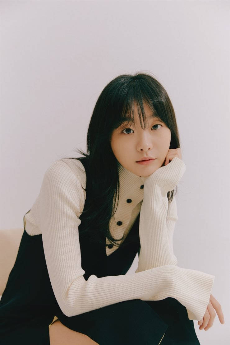 年輕女子身穿米色毛衣和黑色背心，手托下巴擺姿勢。