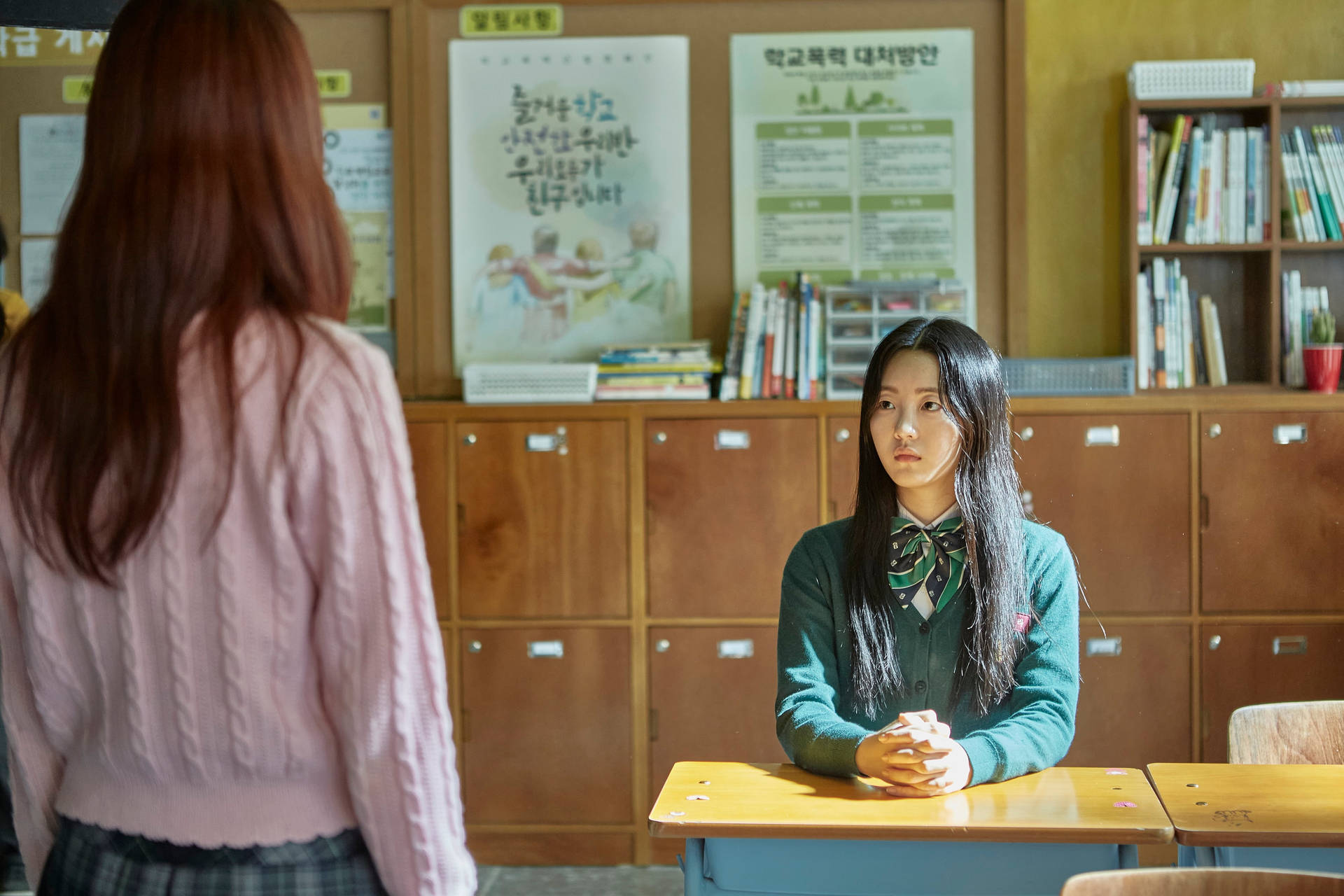 兩個女孩在學校教室裡，一個坐在書桌前抬頭看著另一個背對鏡頭站著的女孩。