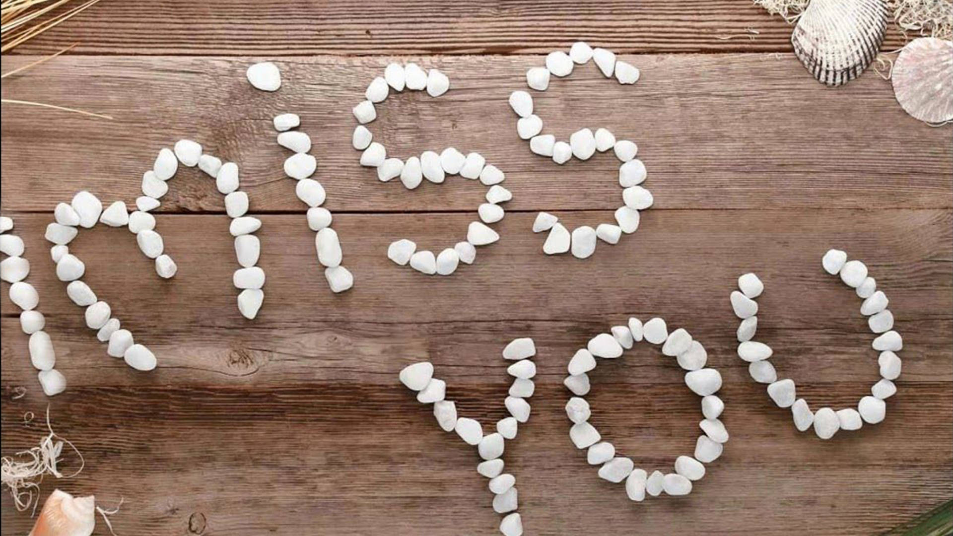 我好想你，石頭 壁紙 — 白色鵝卵石在風化的木質表面上拼出「miss you」，旁邊有貝殼和乾草。