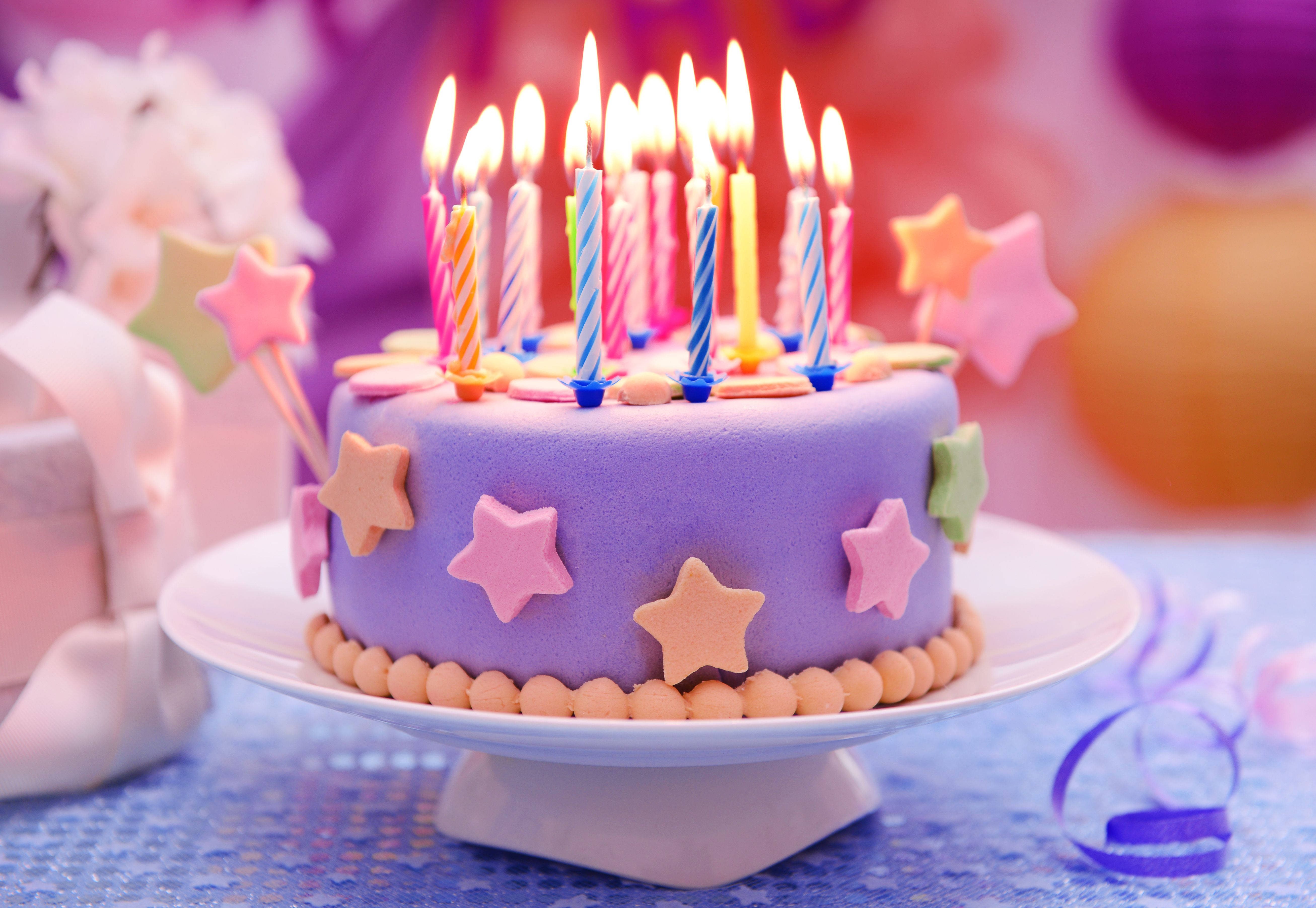 我的生日星星蛋糕 壁紙 — 一個充滿活力的紫色生日蛋糕，上面裝飾著點燃的蠟燭和色彩繽紛的星星裝飾，放在底座上，背景是節日模糊的。