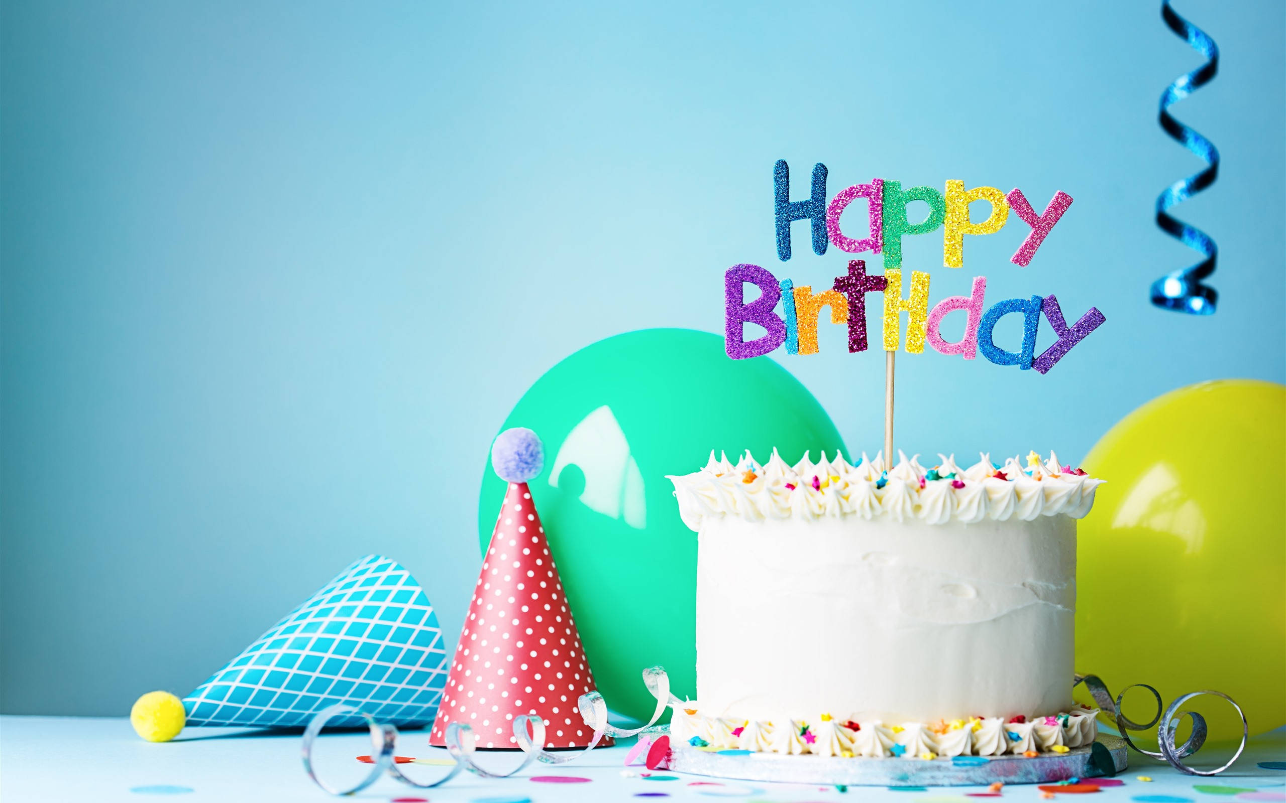 我的生日白色糖珠蛋糕 壁紙 — 一個白色生日蛋糕，上面有彩色糖粒和閃亮的「Happy Birthday」裝飾，旁邊有派對帽和氣球，背景為藍色。