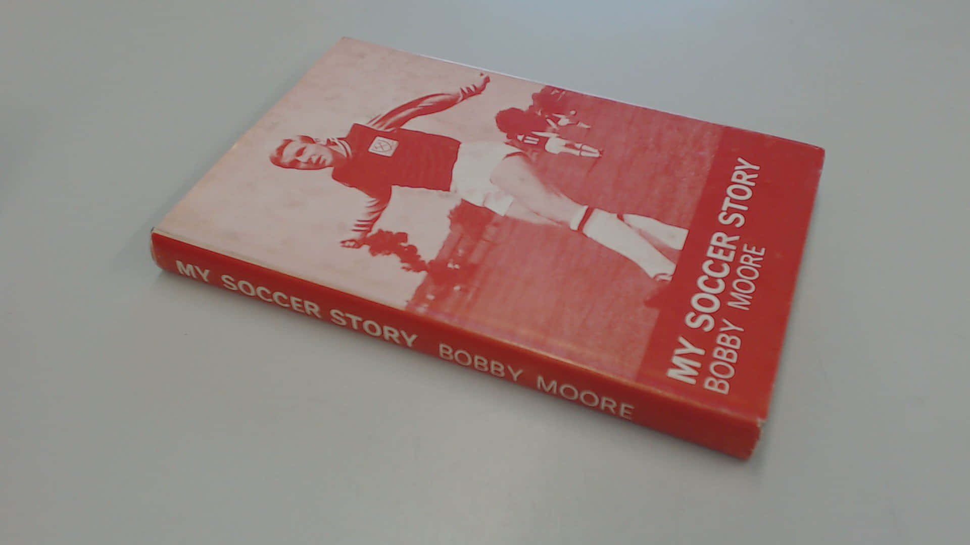 我的足球故事 鮑比·穆爾 壁紙 — 一本紅色書，名為《My Soccer Story》，作者Bobby Moore，傾斜放在淺灰色平面上。