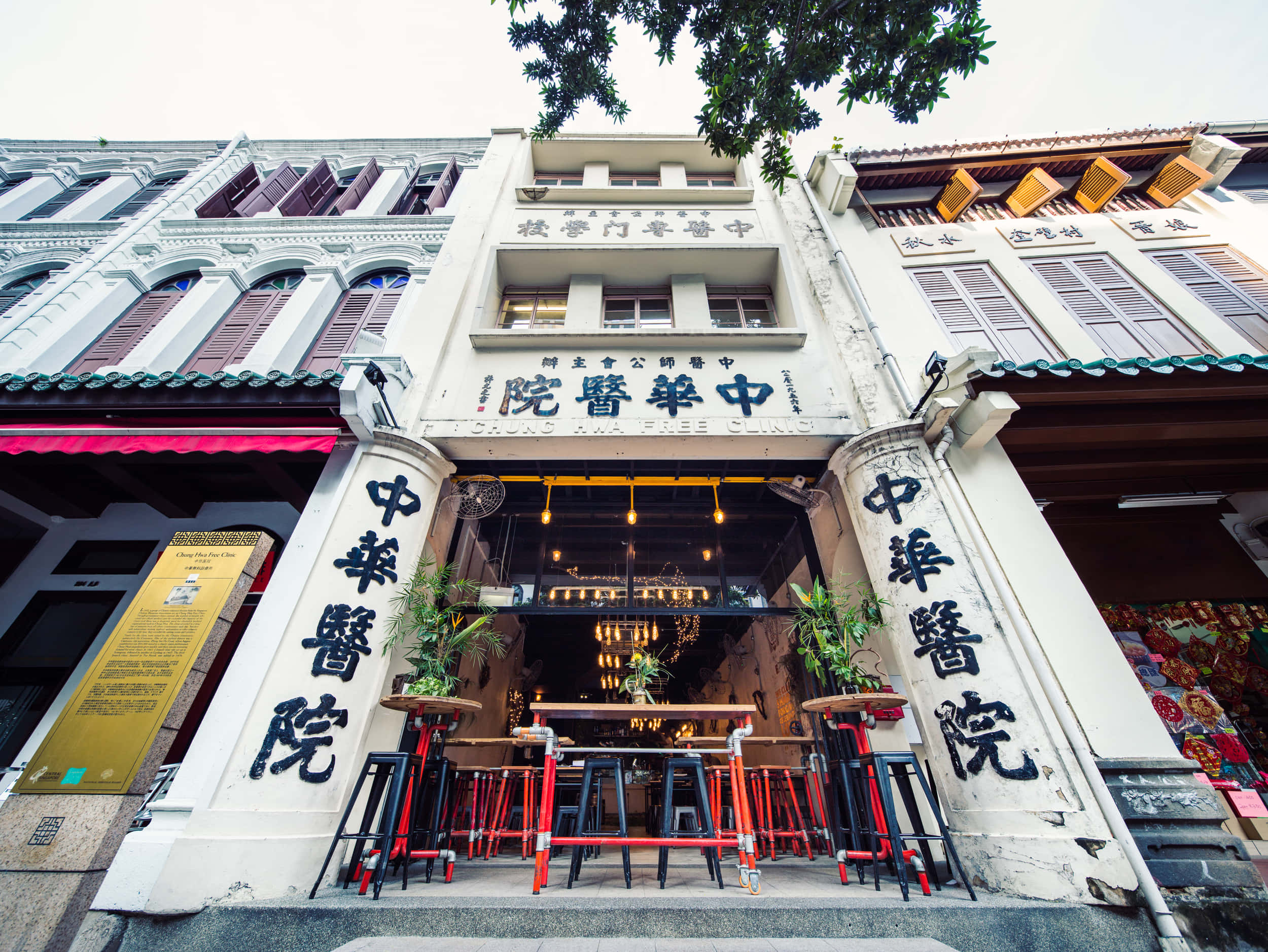 我的酷炫咖啡館圖像 壁紙 — 充滿活力的街頭景觀，歷史悠久的店屋柱子上有中文字，現代酒吧外有紅色凳子。