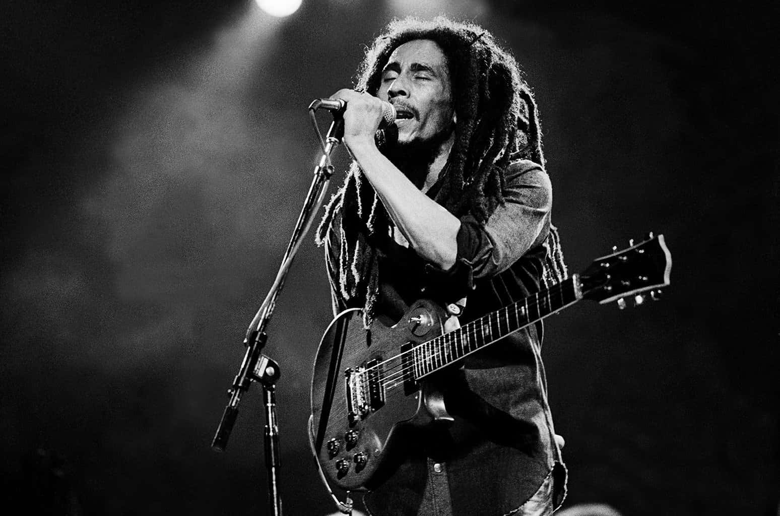 「我的音樂是我的抗議之聲」- 鮑勃·馬利 壁紙 — Bob Marley 在黑白舞台燈光下，熱情地對著咪高峰唱歌，同時手持結他。