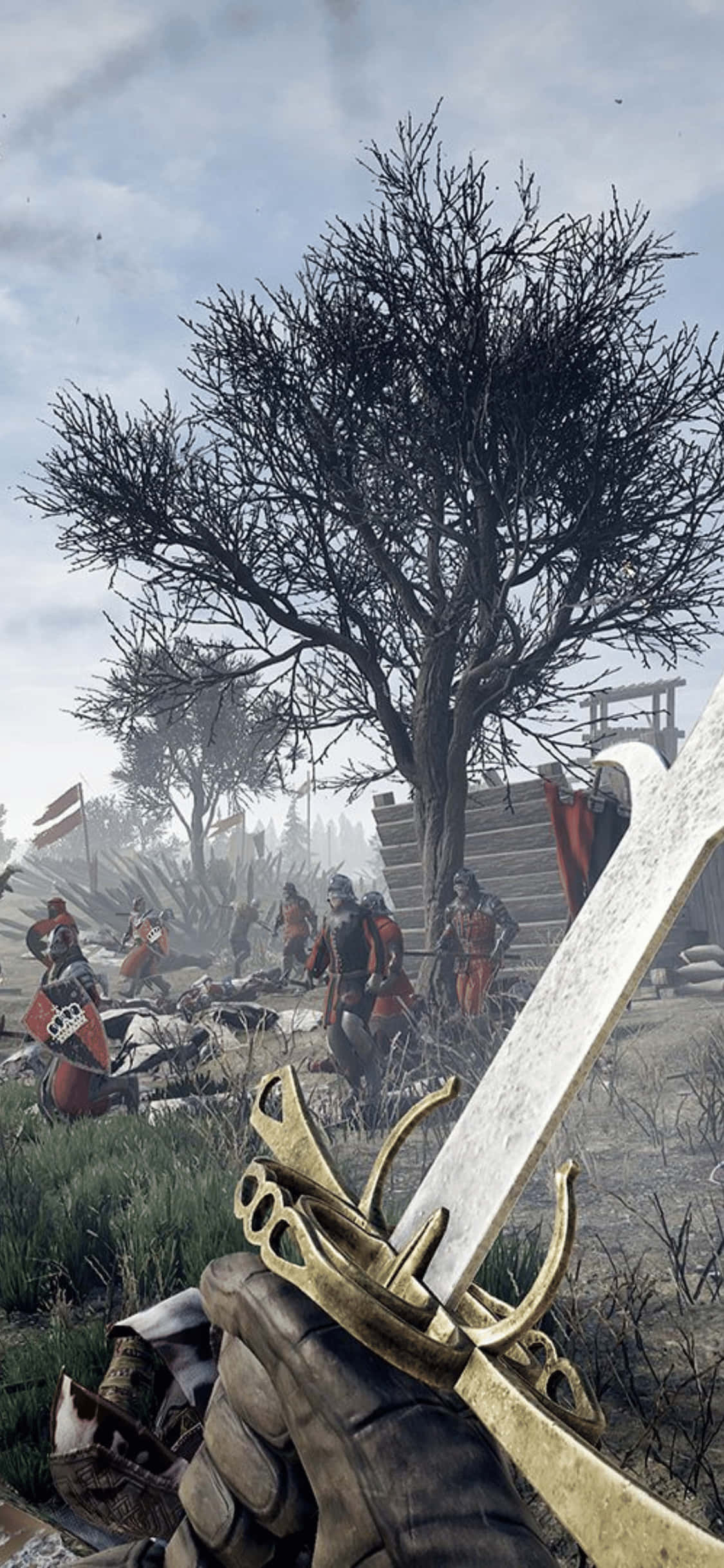 戰士戰場 iPhone XS Mordhau 背景 壁紙 — 第一人稱視角，戴手套的手握著一把裝飾華麗的金色劍柄的劍，面對著一個有士兵和一棵光禿禿的樹的戰場，天空陰沉。