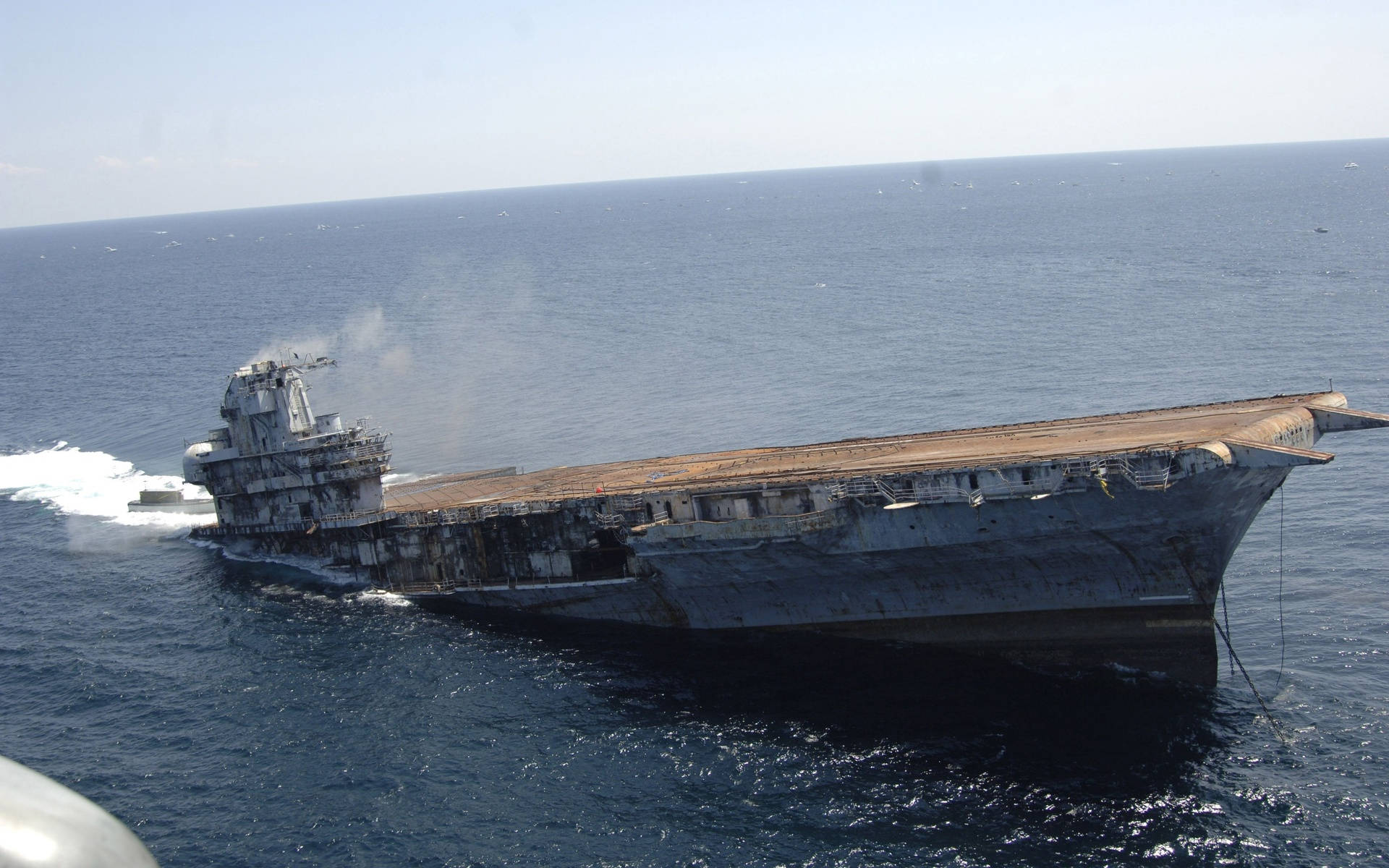 戰艦沉沒於海洋 壁紙 — 一艘生鏽的航空母艦在清澈的天空下傾斜在深藍色的海洋中，甲板上冒著煙。
