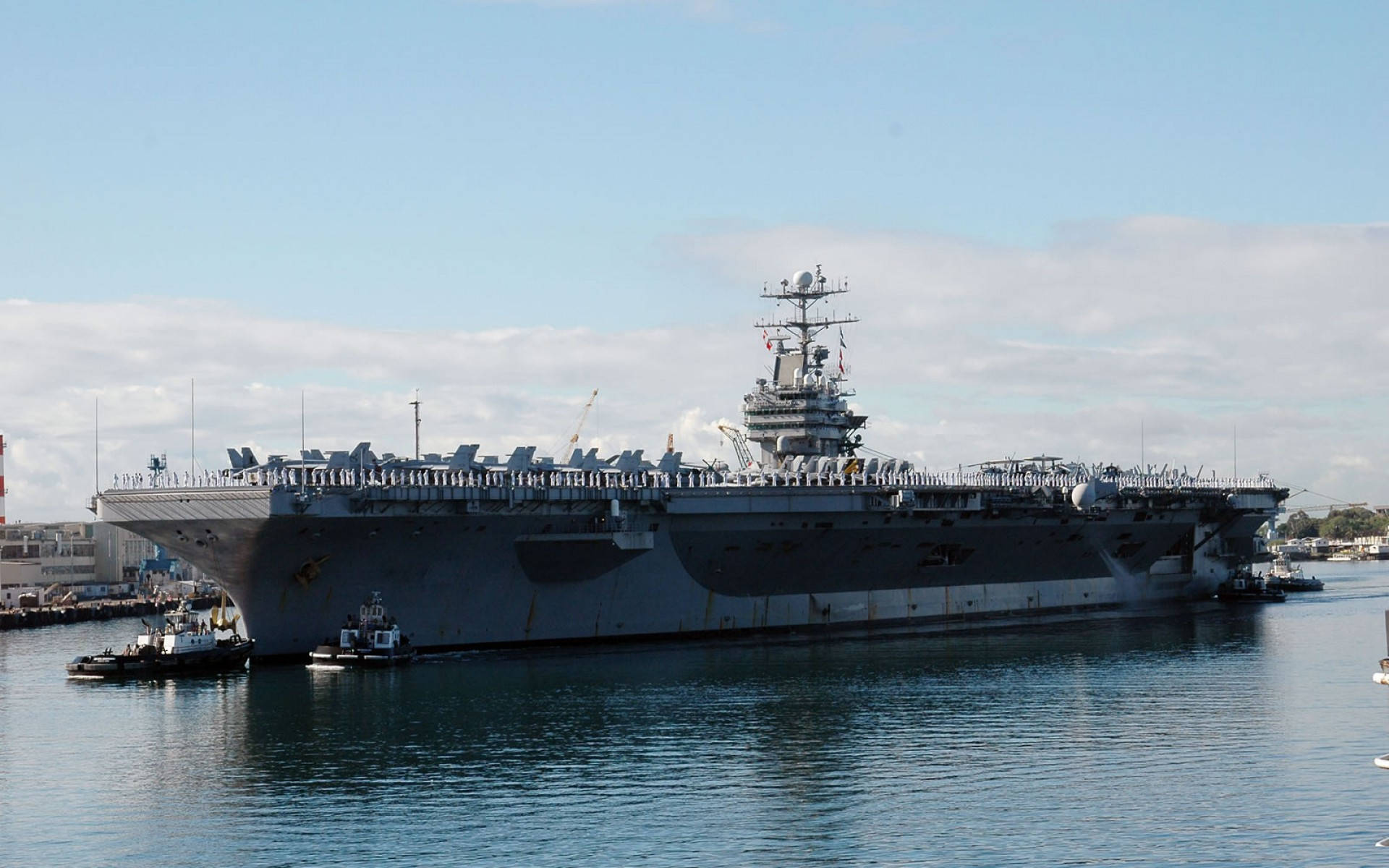 戰艦由小艇護航 壁紙 — 一艘巨大的灰色航空母艦停泊在平靜的港灣水域中，附近有小拖船，天空部分多雲。