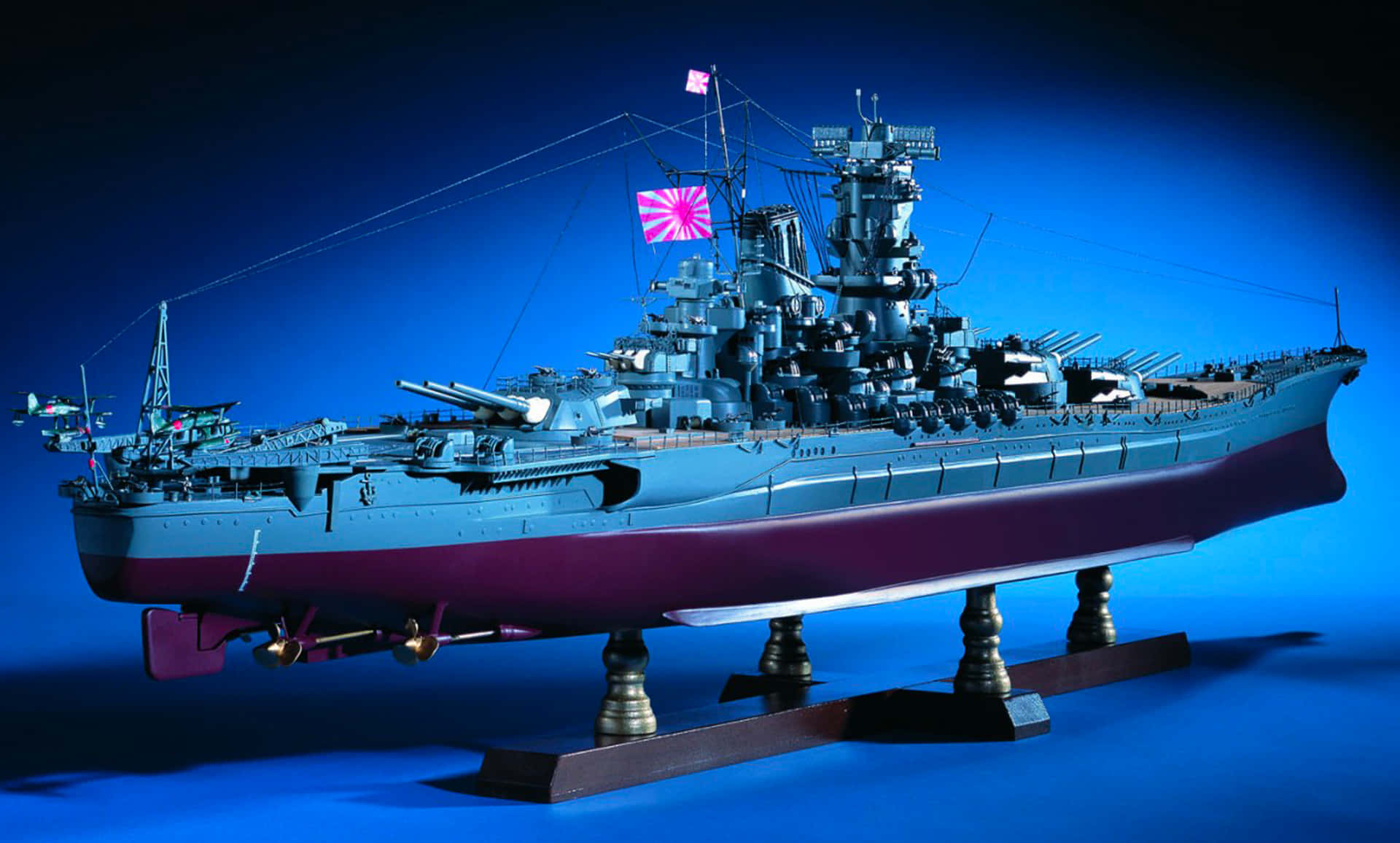 戰艦的火力威懾 壁紙 — 一艘帶有紅白旗的日本戰艦精細比例模型，矗立在展示架上，背景為深藍色。