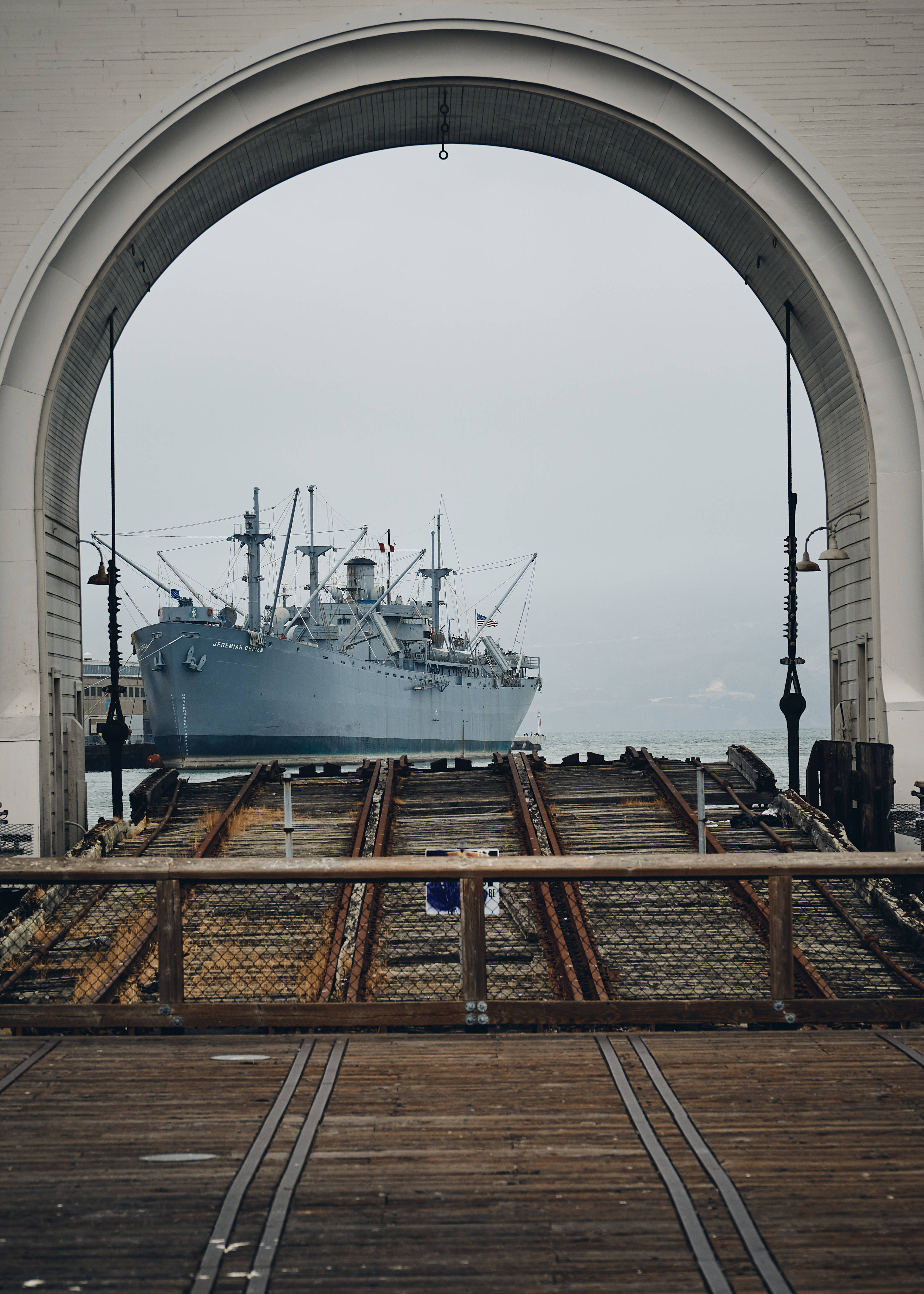 戰艦穿過拱門 壁紙 — 一艘灰色大船停泊在經過風化的木製斜坡之外，斜坡由白色拱形結構框住，天空多雲。