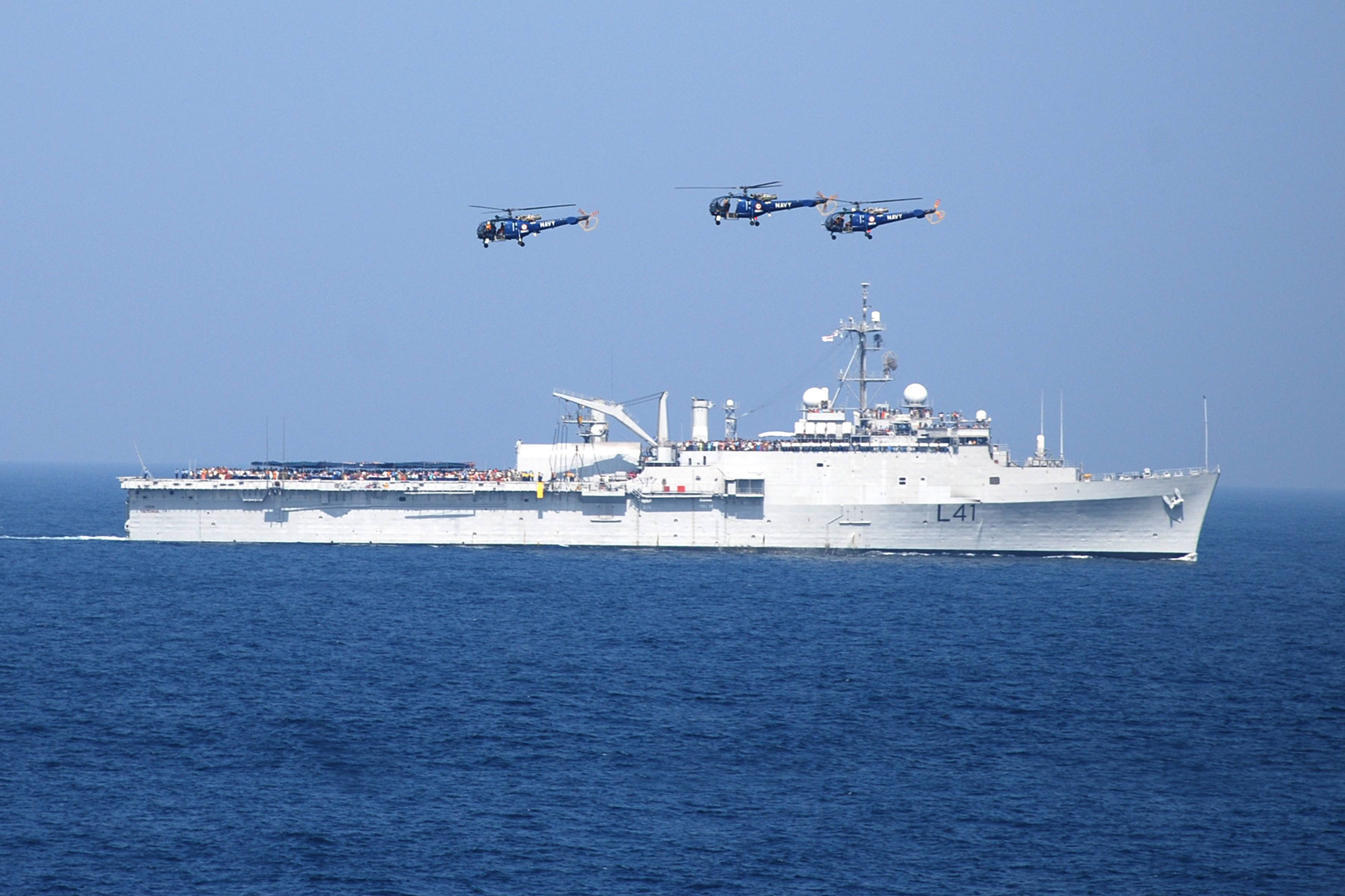 戰艦與懸浮直升機 壁紙 — 一艘大型灰色軍艦在深藍色海洋上航行，三架直升機在晴朗天空下以編隊飛行。