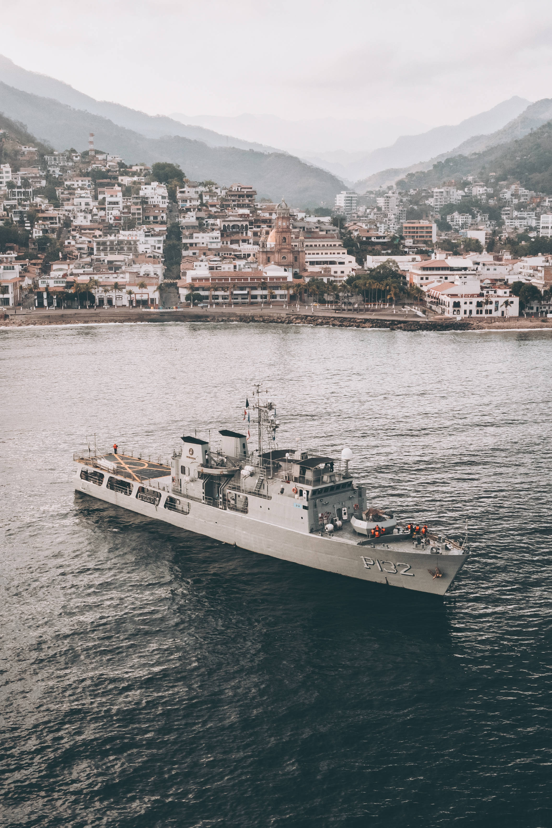 戰艦與擁擠城市 壁紙 — 一艘灰色海軍艦艇在黑暗水域中平靜航行，背景是沿海小鎮和霧氣繚繞的山脈。