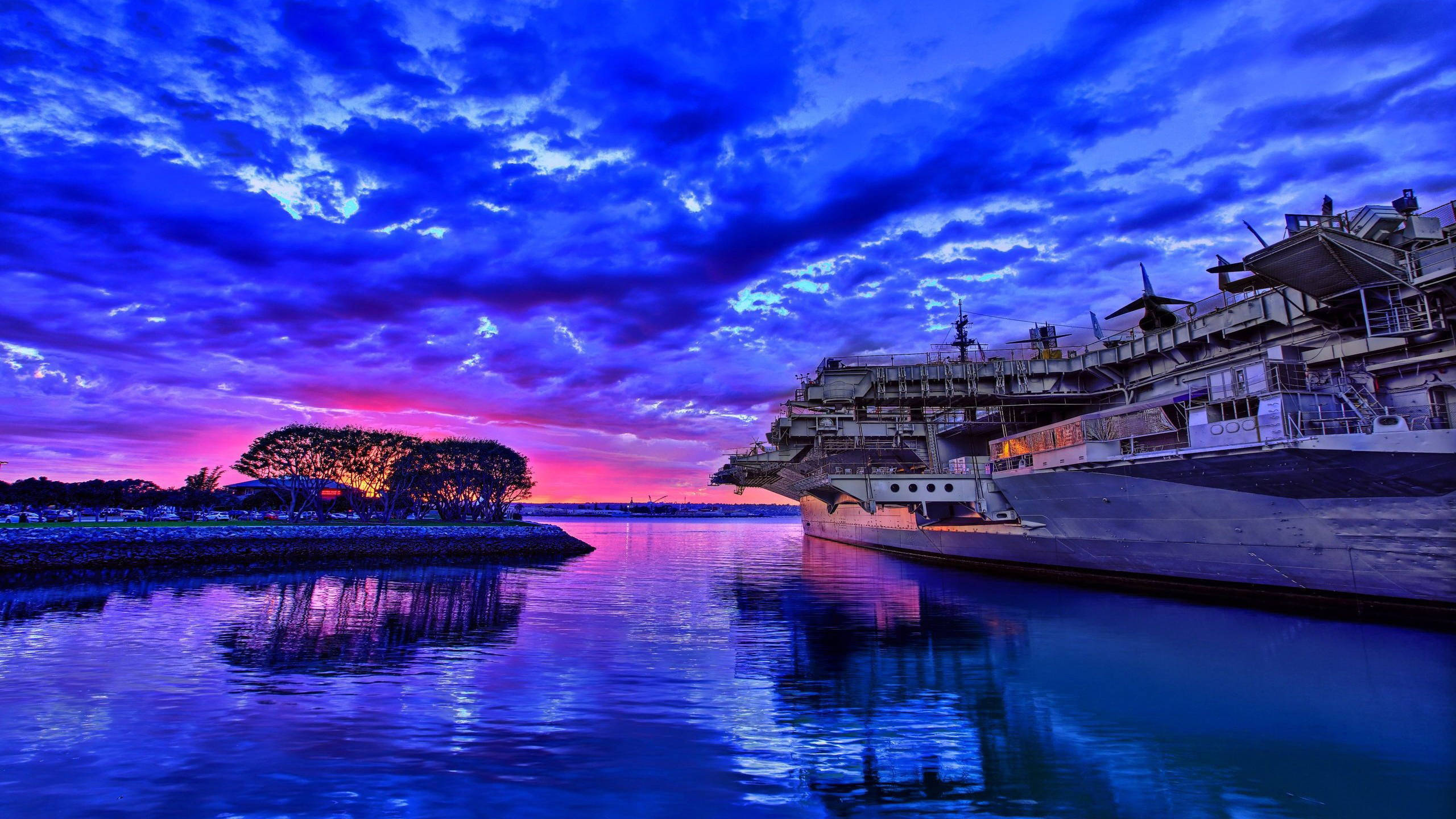 戰艦與藍天雲朵 壁紙 — 一艘巨大的航空母艦在平靜的水域中停泊，天空呈現戲劇性的暮色，塗滿了電光藍和火熱粉紅。