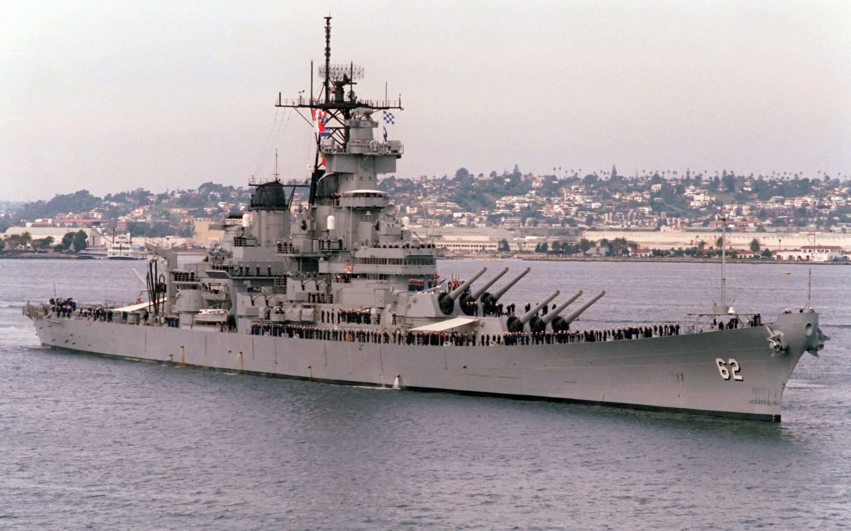 戰艦艏部駛過開闊海域 壁紙 — 一艘巨大的灰色戰艦，編號62，在朦朧的沿海城市背景下平靜地航行。