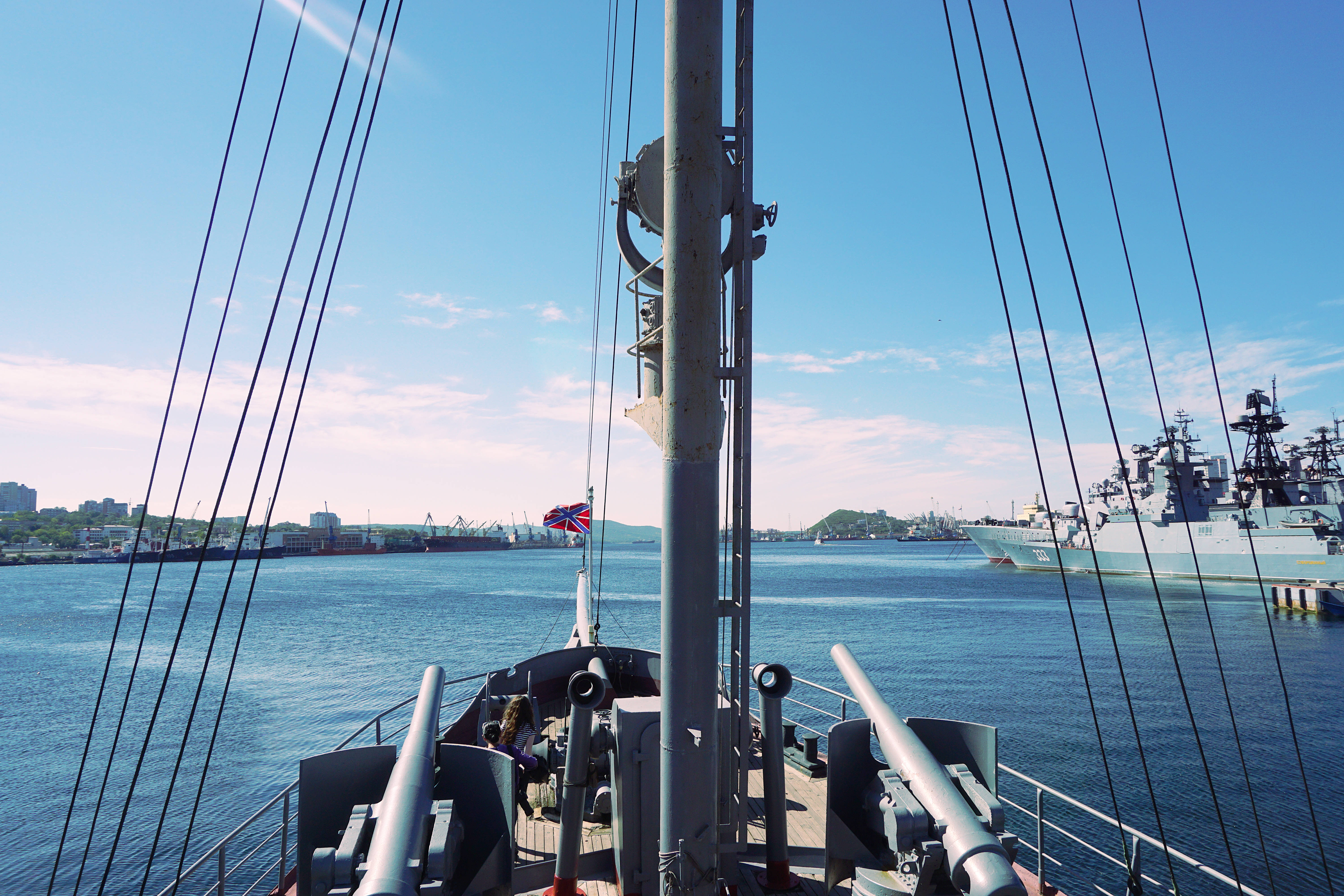 戰艦觀景甲板 壁紙 — 從歷史戰艦甲板上的景色，展示大炮、索具和英國國旗，以明亮的藍天為背景，港口內有其他海軍艦艇。