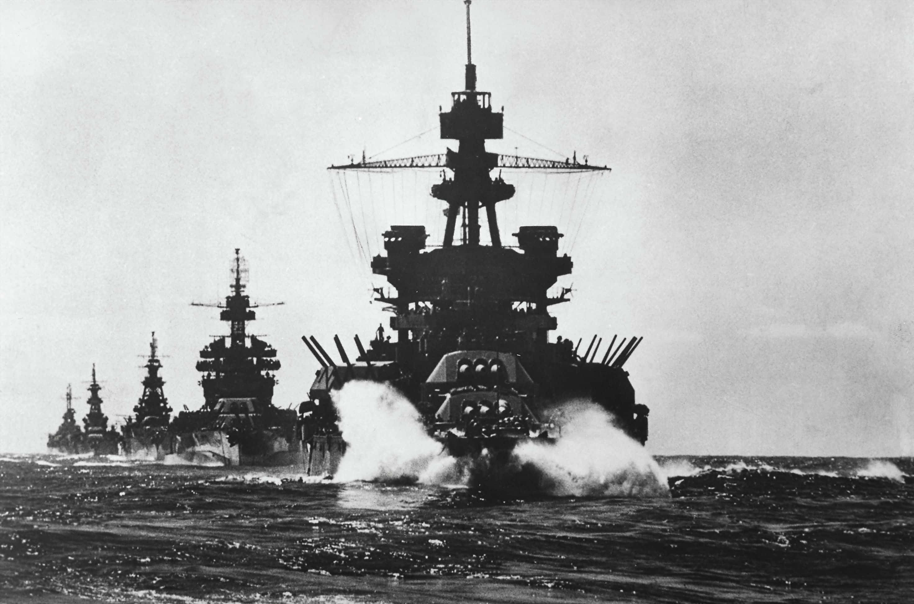 戰艦駛入戰鬥熱點 壁紙 — 一排巨型戰艦劃過波濤洶湧的海面，在陰天留下白色航跡。