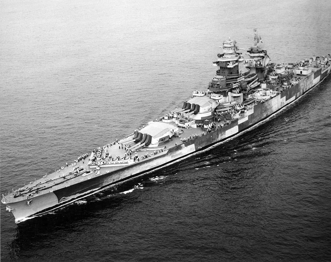 戰艦駛入港灣 壁紙 — 一艘巨大的戰艦在平靜的海面上航行，甲板上可見船員，呈現出引人注目的黑白航拍視角。
