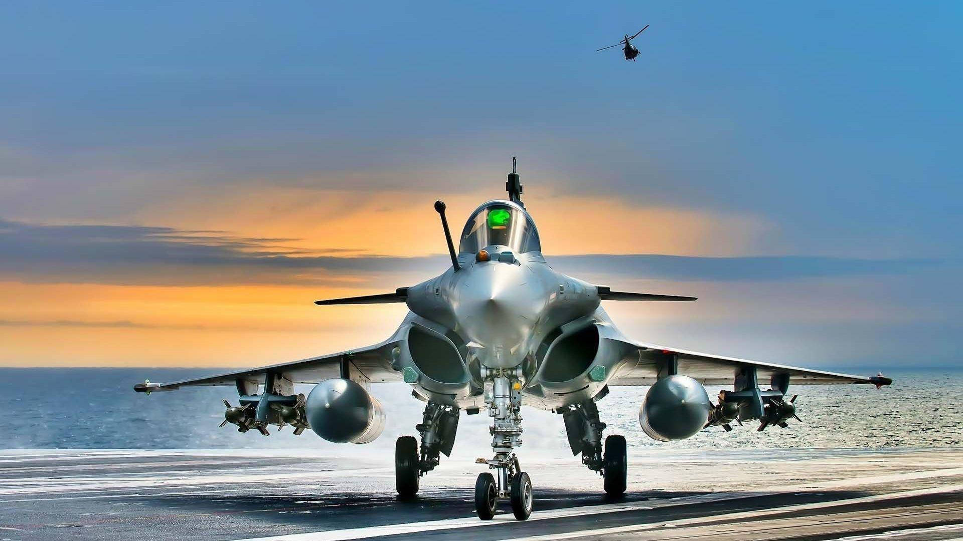 戰鬥機飛越天空的空中視角 壁紙 — 一架流線型戰鬥機在日落時分在航空母艦甲板上滑行，一架直升機在上空飛行。