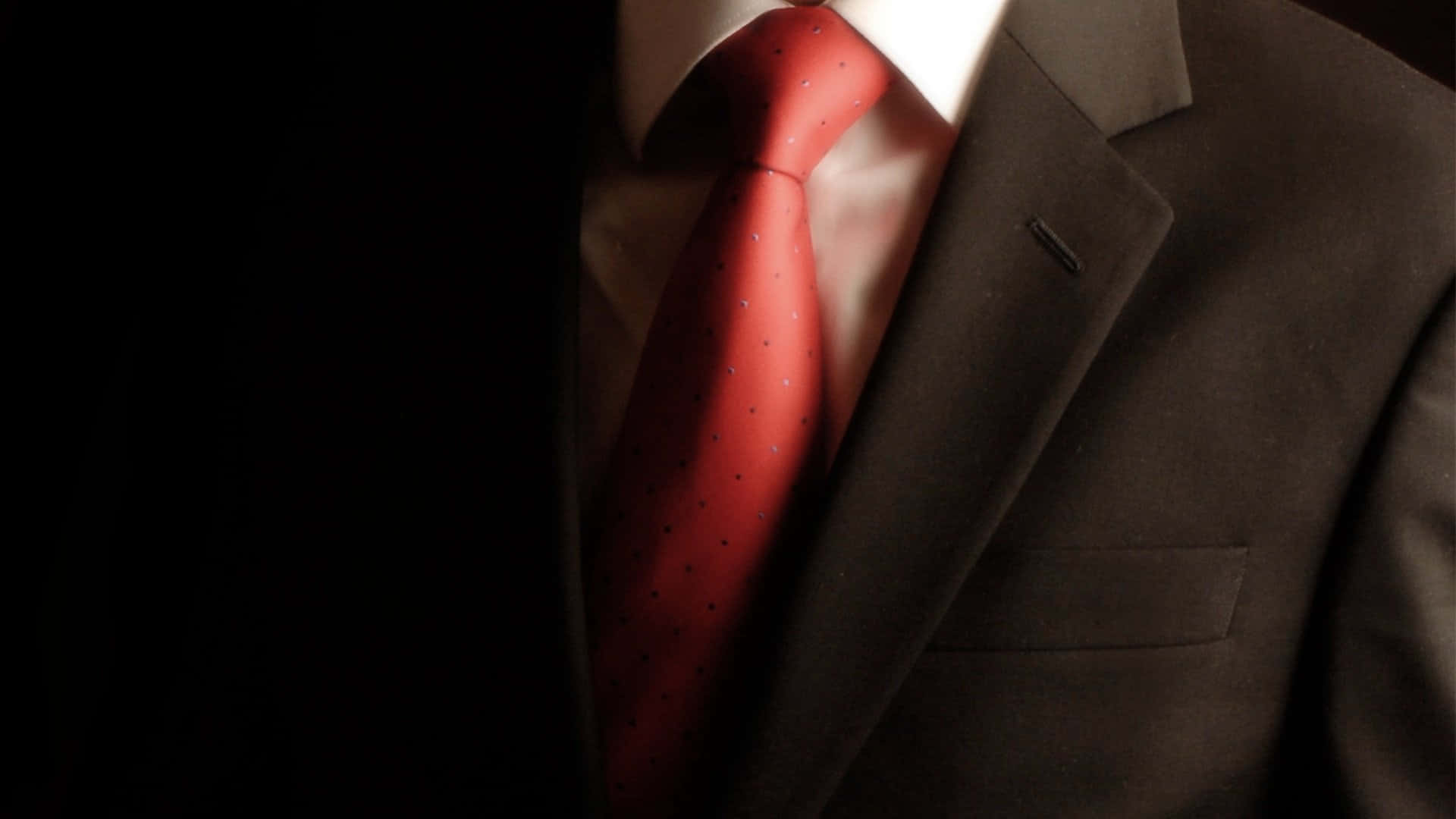 Red Tie 桌布 壁紙 — 戲劇性燈光下，一位衣著講究的男士上半身特寫，深色西裝與白襯衫搭配紅色圓點領帶。