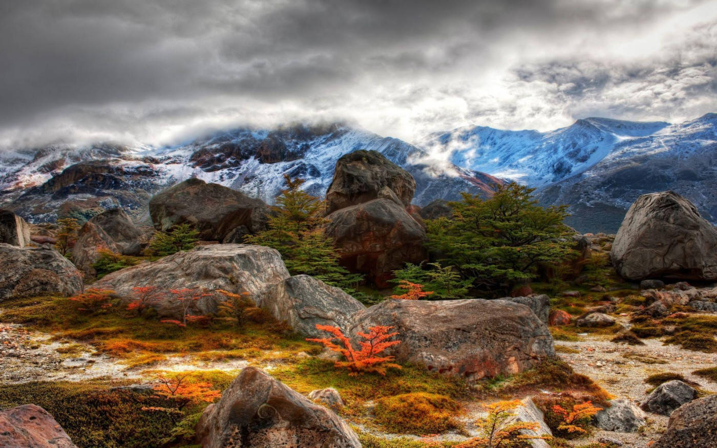 Mountain 桌布 壁紙 — 戲劇性的山景，有白雪覆蓋的山峰、巨大的岩石和充滿活力的橙色樹葉，在陰天天空下。