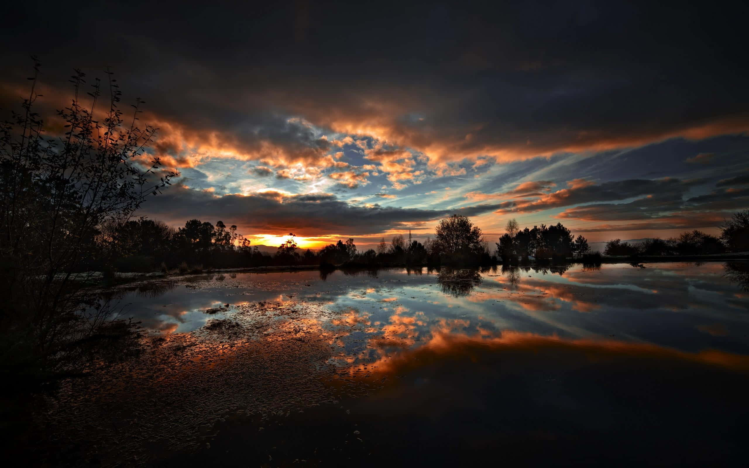 Sunset Reflection 桌布 壁紙 — 戲劇性的日落描繪出火橙色和深藍色的雲彩，完美地倒映在一個被剪影樹木環繞的平靜黑暗池塘中。