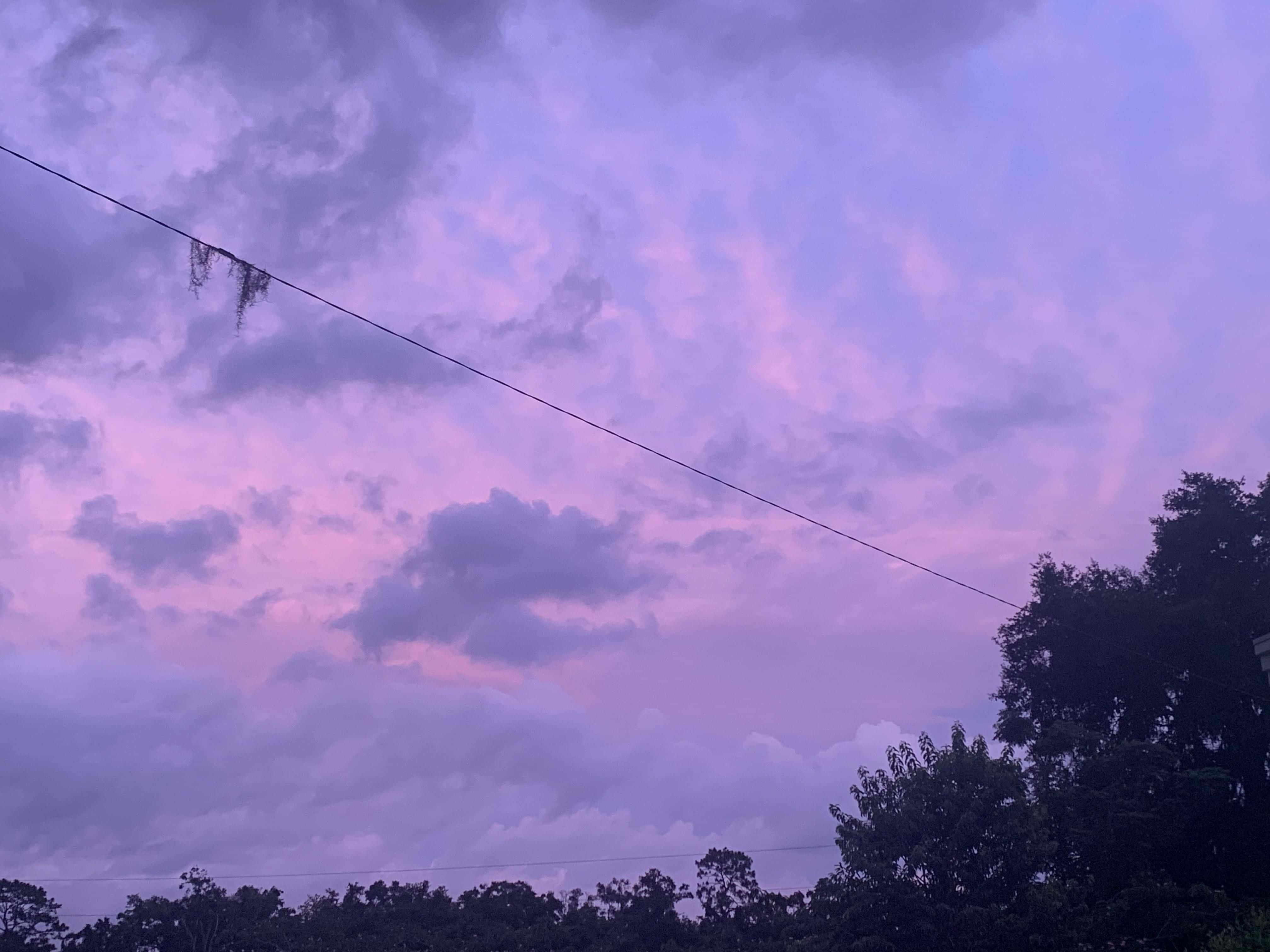 唯美雲朵 桌布 壁紙 — 戲劇性的暮色天空，以柔和的紫色和粉紅色繪製，下方有深色雲朵和樹木剪影。