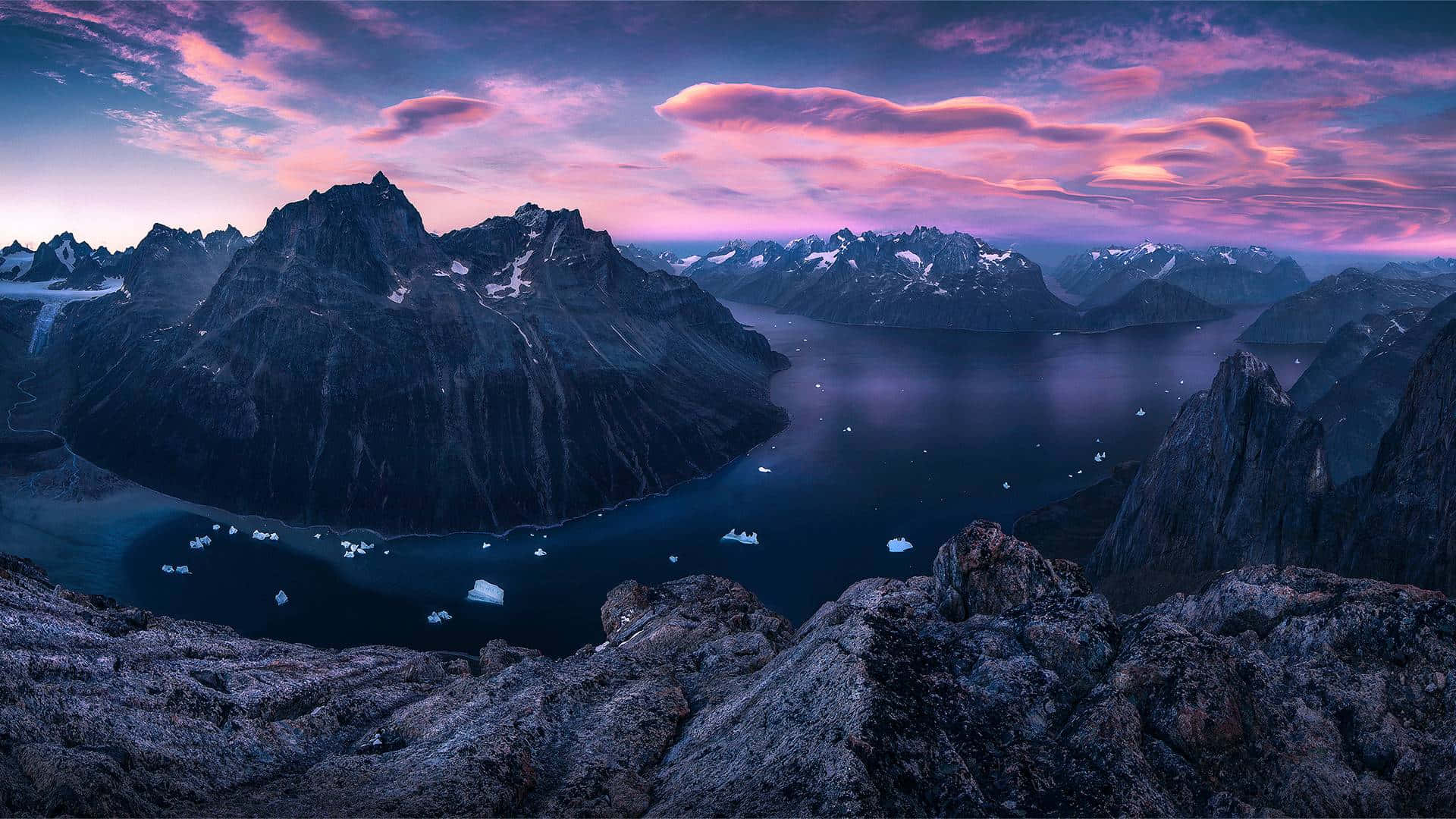 Greenland 桌布 壁紙 — 戲劇性的暮色景觀，崎嶇的山脈環繞著深藍色的峽灣，點綴著漂浮的冰山，天空呈現充滿活力的粉紅色和紫色。