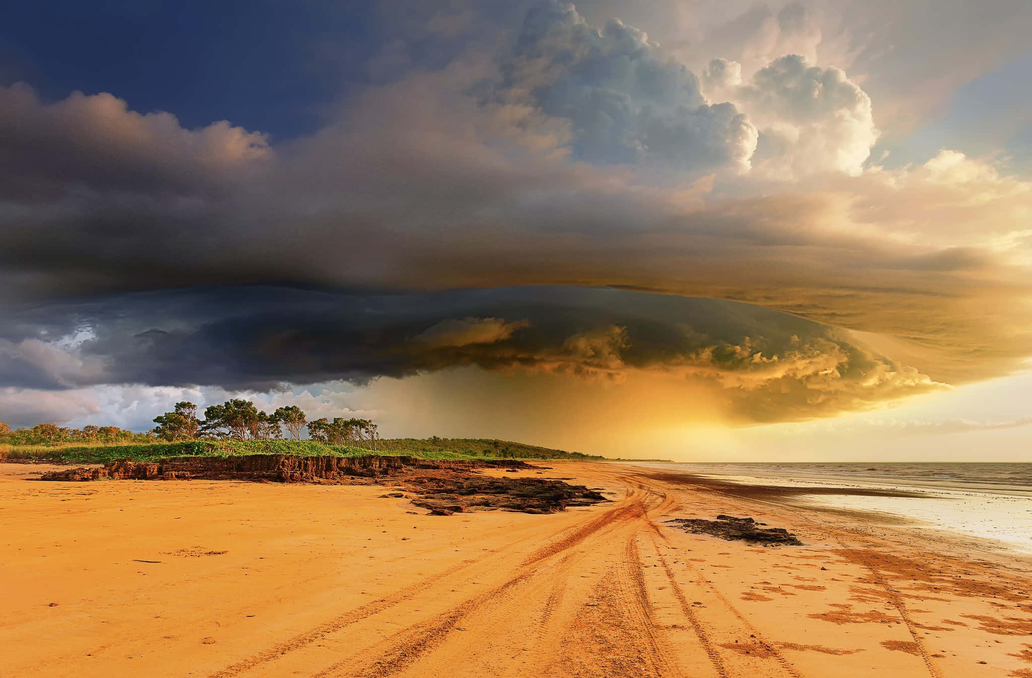 Seashore 桌布 壁紙 — 戲劇性的暴風雲籠罩在日落時分的金色沙灘上，將溫暖的光線投射在濕沙和遠處的樹木上。
