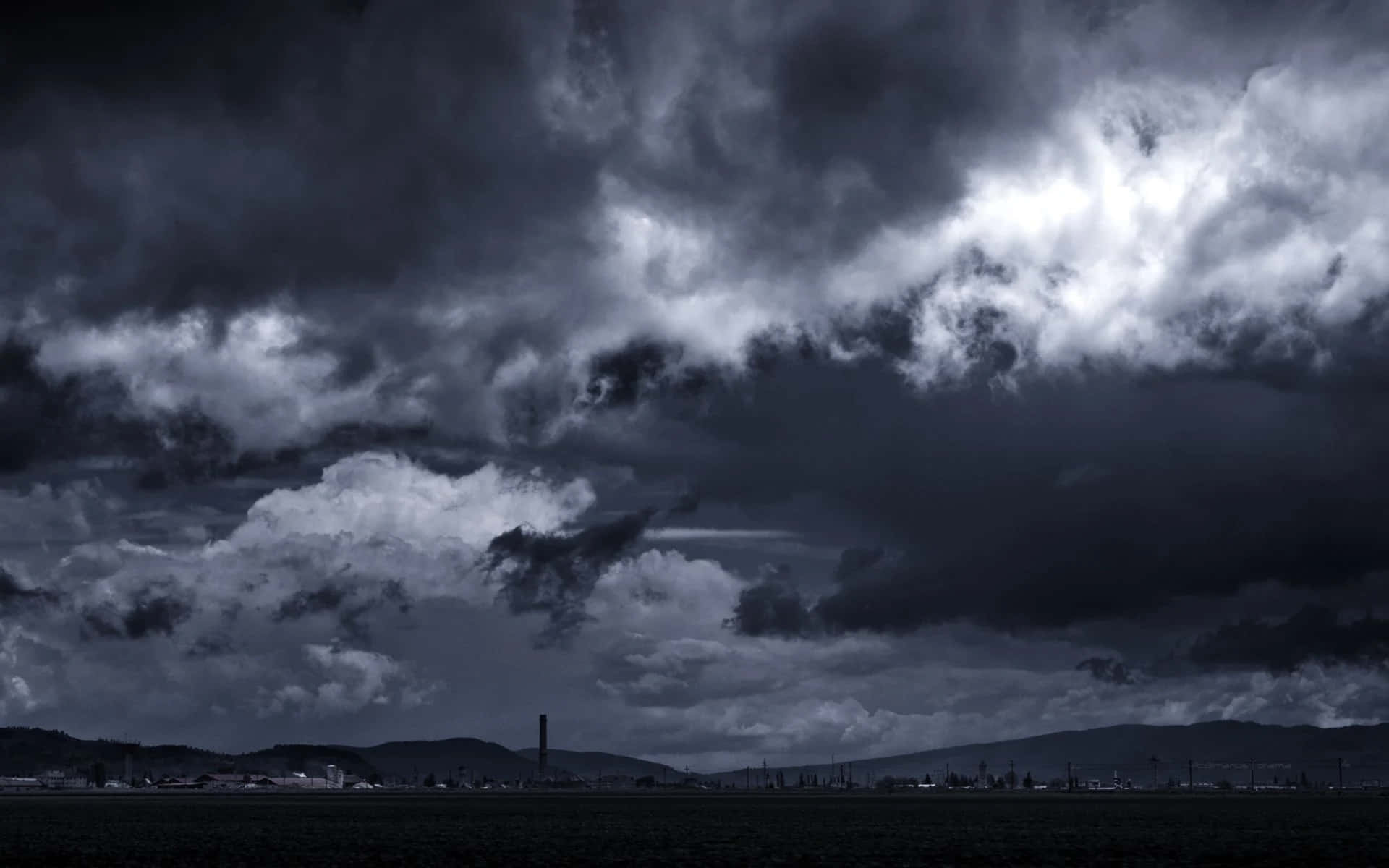 Black And White Storm 桌布 壁紙 — 戲劇性的暴風雲籠罩著昏暗的鄉村景觀，遠處的山丘和模糊的工業建築在陰鬱的暮色天空下若隱若現。