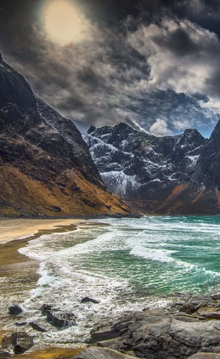 Norway 桌布 壁紙 — 戲劇性的海岸場景，在暴風雨的天空下，高聳的雪山下的沙灘上，碧綠色的海浪拍打著。
