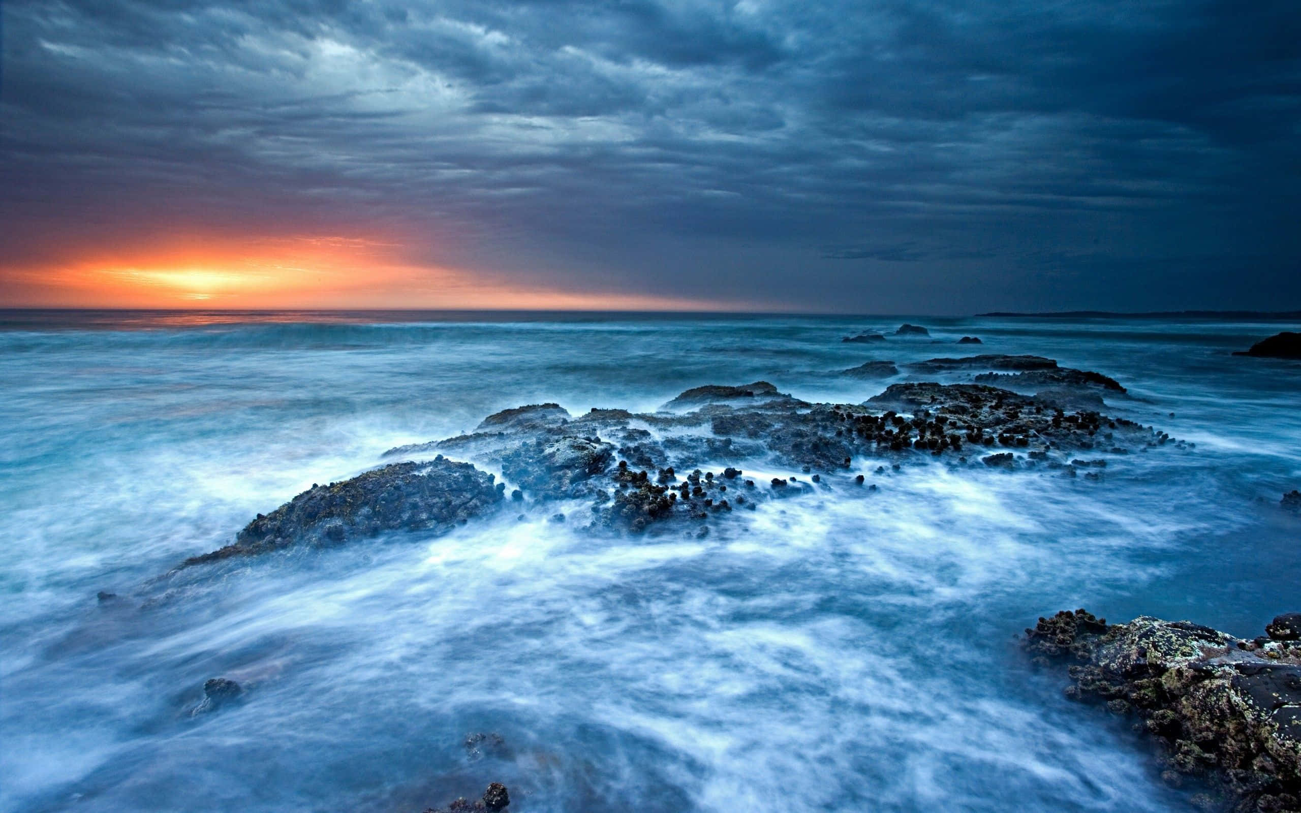 海浪 桌布 壁紙 — 戲劇性的海浪在暴風雨的天空下衝擊著黑暗的岩石，地平線上燃燒著夕陽的餘暉。