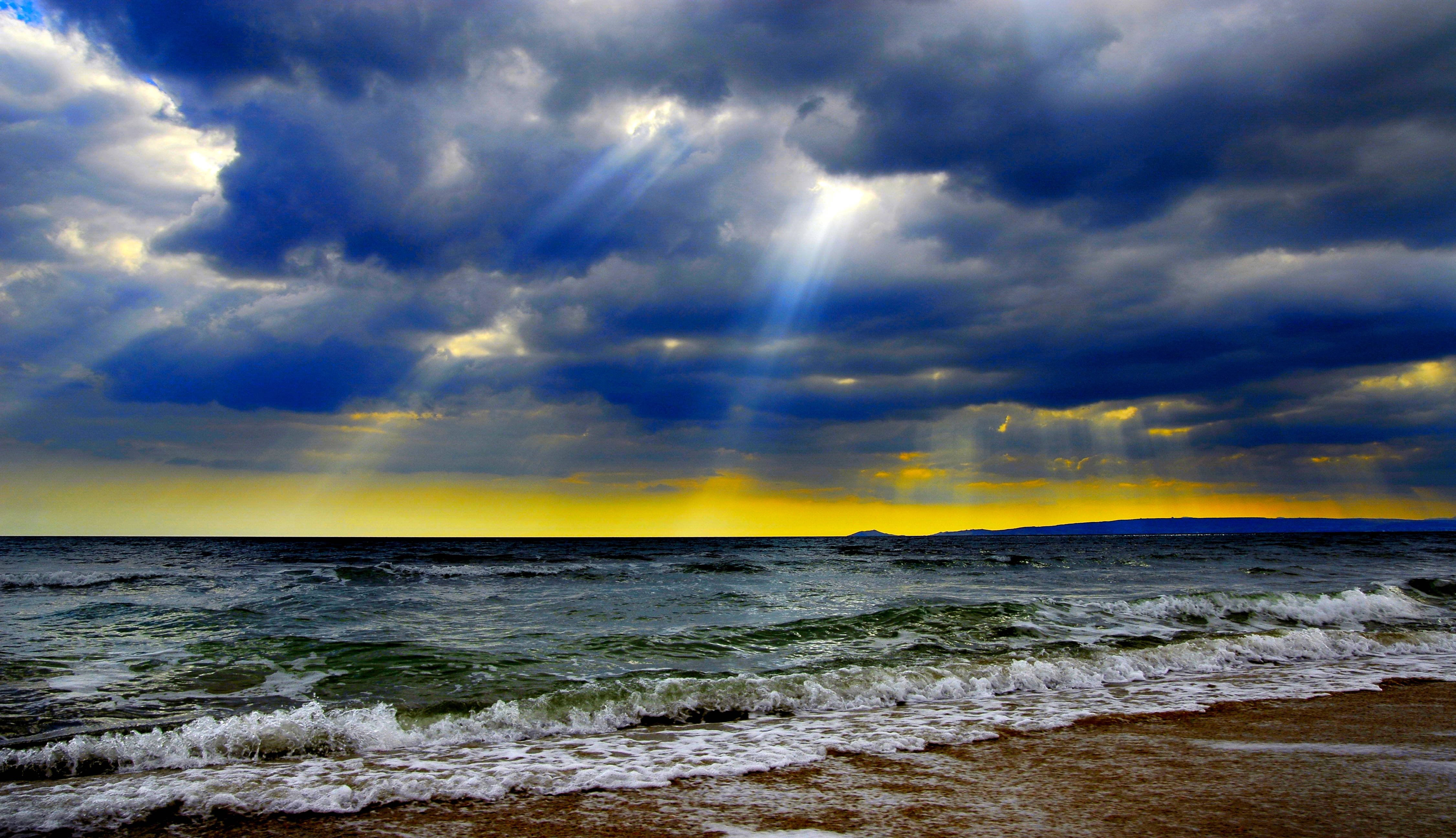 Sun Rays 桌布 壁紙 — 戲劇性的陽光穿透黑暗的暴風雲，照耀在波濤洶湧的大海與沙灘交匯處。