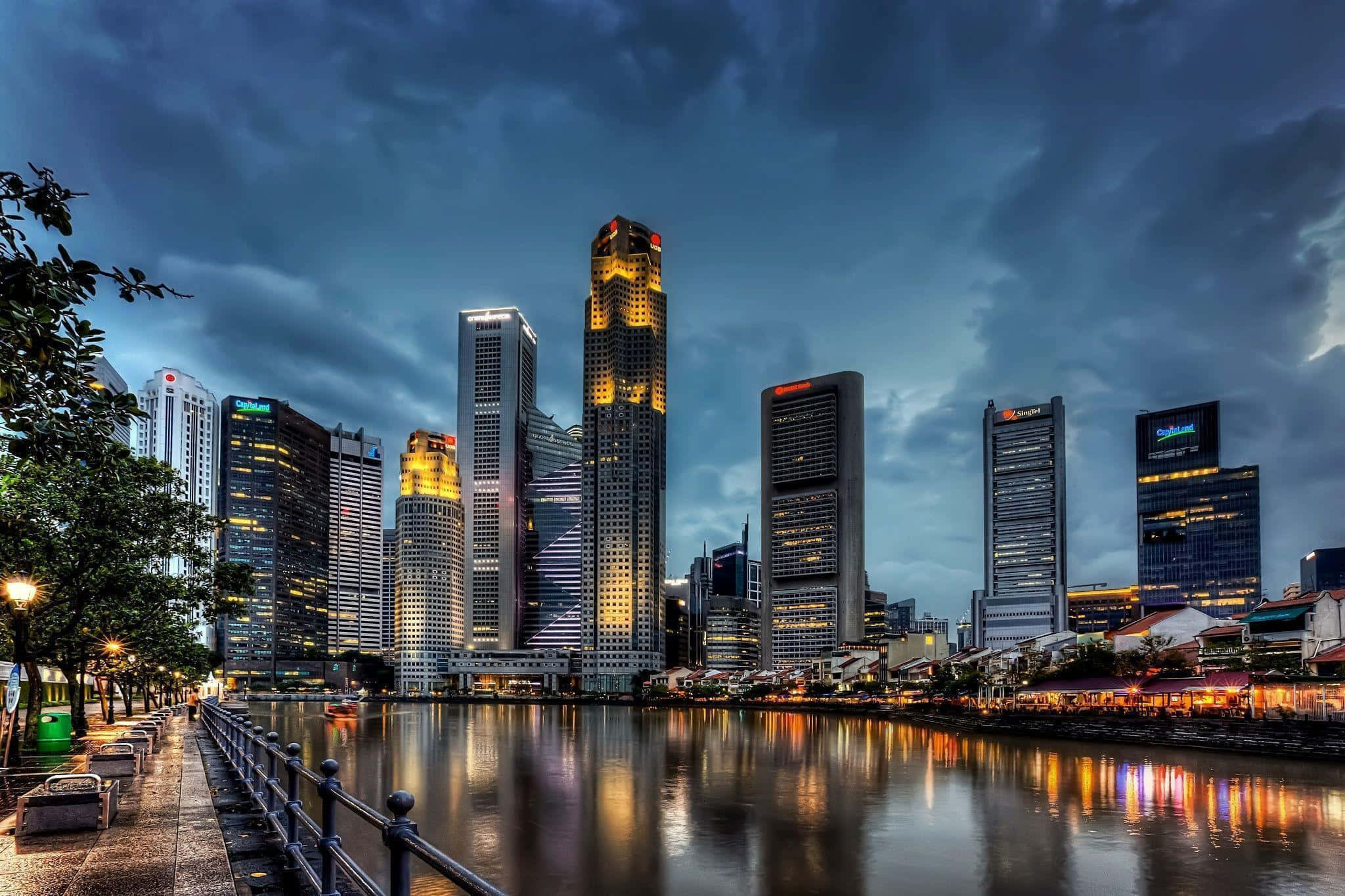 Singapore 桌布 壁紙 — 戲劇性的黃昏城市景觀，高聳的摩天大樓在平靜的河面上反射出溫暖的燈光，天空陰沉。