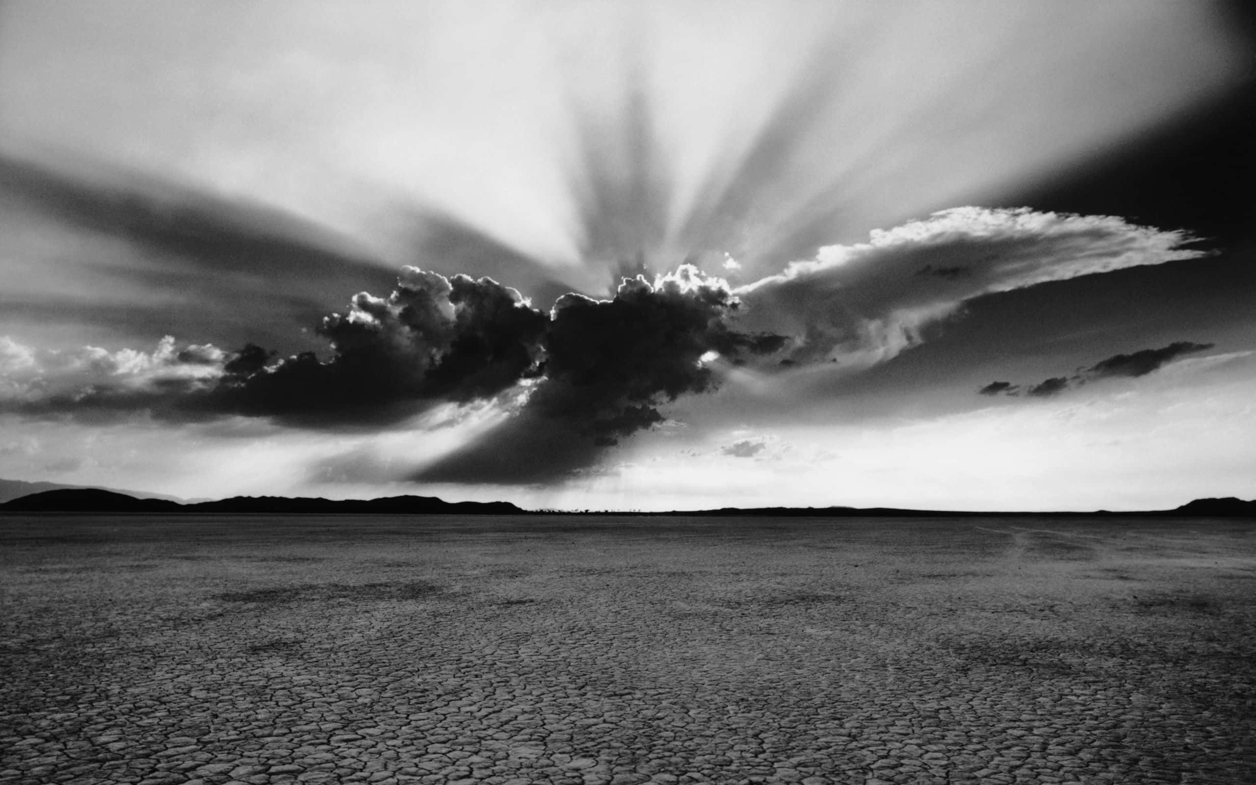 Black And White Sky 桌布 壁紙 — 戲劇性的黑白照片，陽光穿過暴風雲層照射在龜裂的沙漠平原上。