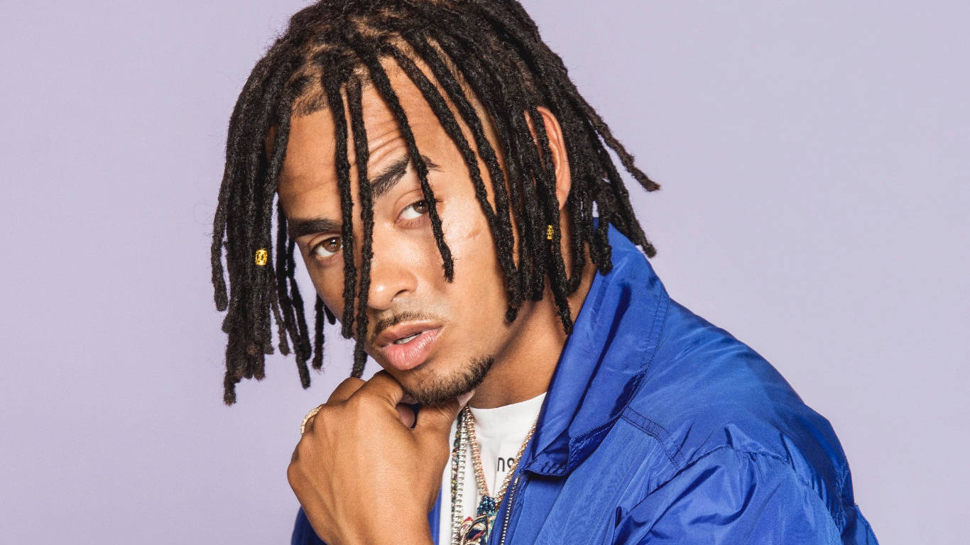 戴 dreadlocks 的歌手奧祖納 壁紙 — 一位年輕男子留著長辮子，戴著金耳環，身穿藍色外套，在柔和的薰衣草背景前自信地擺姿勢。