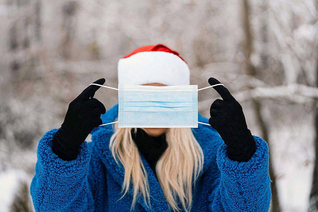 戴口罩女子冬季桌面 壁紙 — 一個穿著藍色外套和聖誕帽的人，在雪白的冬季背景下，將醫用口罩遮在臉上。