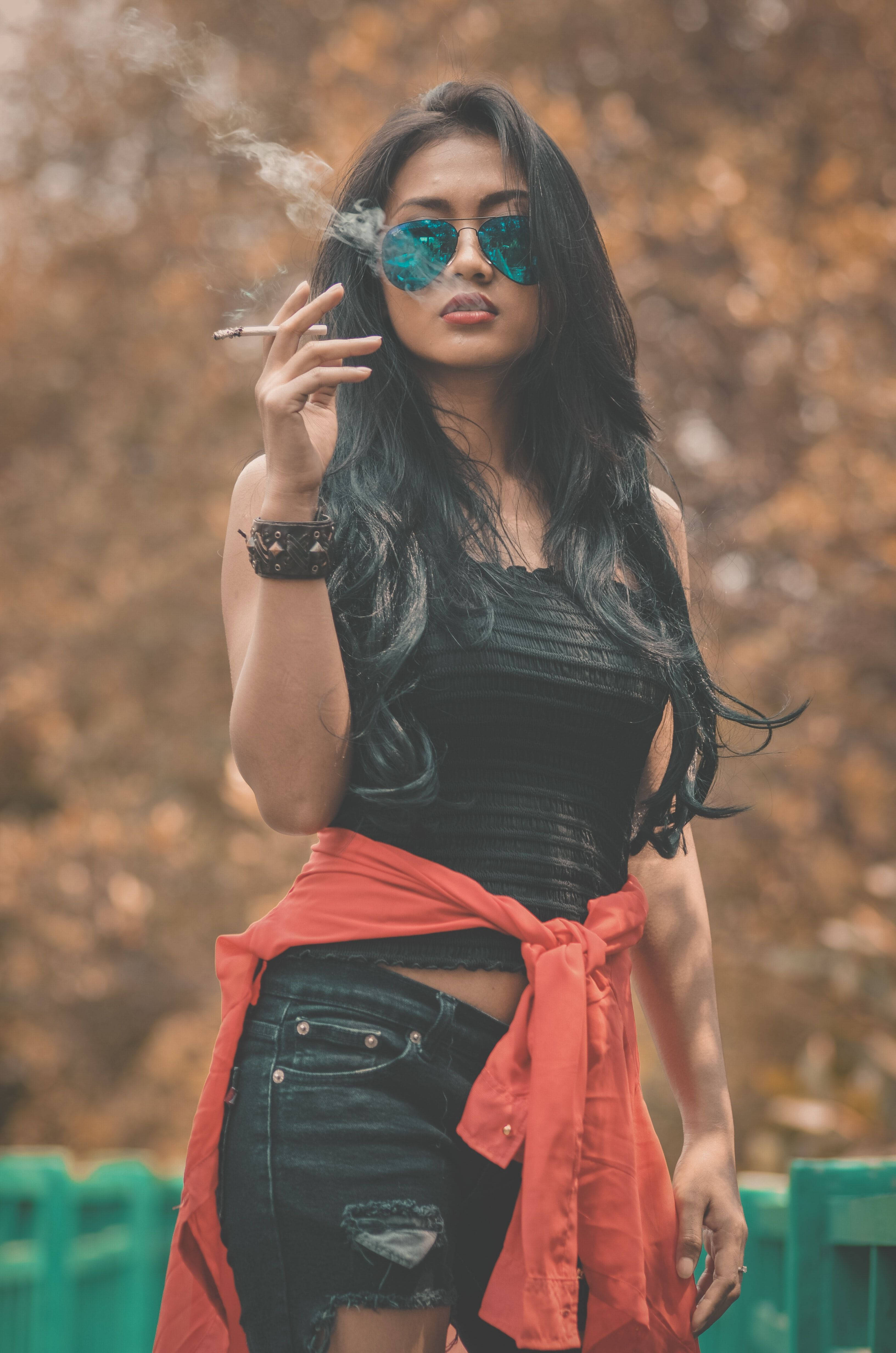 戴太陽眼鏡抽菸的酷態度女孩 壁紙 — 一位時尚的長黑髮女性，戴著反光太陽眼鏡，紅色外套綁在腰間，在模糊的秋日背景下抽煙。