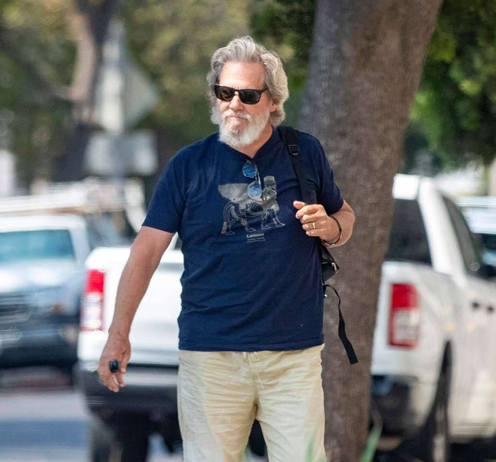 灰髮男子戴著太陽眼鏡走在陽光明媚的街道上，旁邊有停放的汽車。