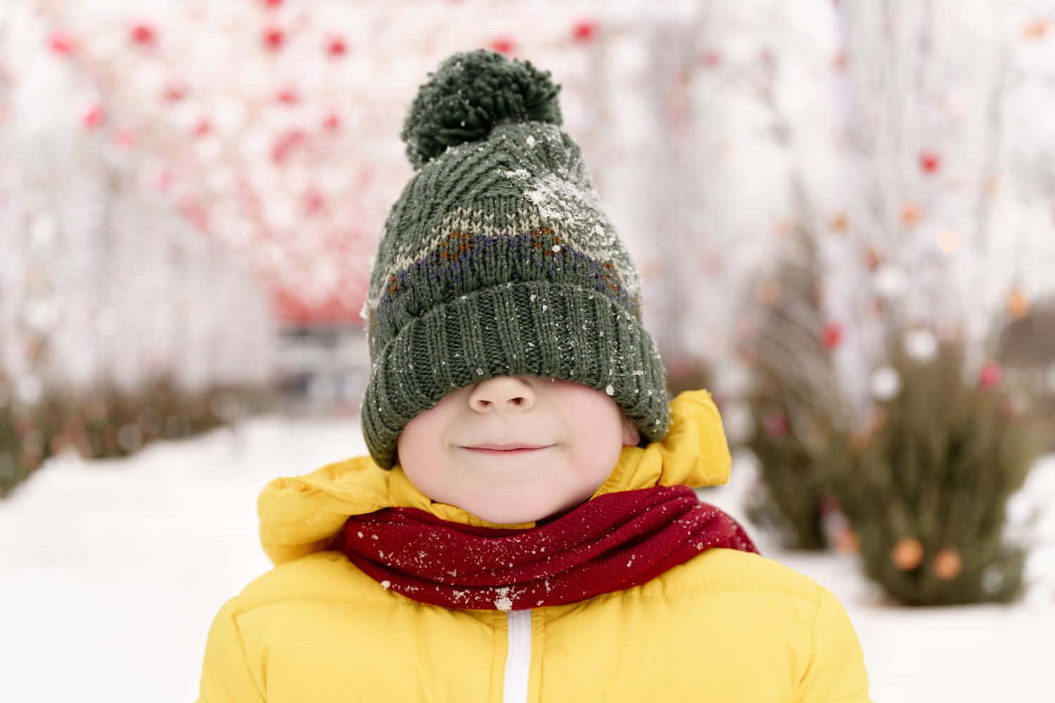 戴著綠色帽子和黃色外套的小孩在雪白的冬日環境中微笑。