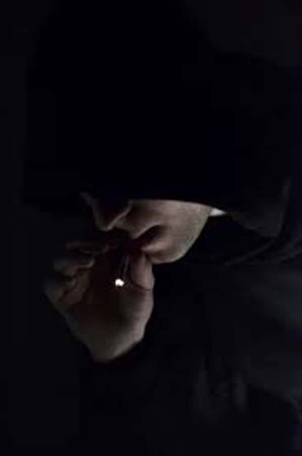 戴帽憂鬱少年抽菸壁紙 壁紙 — 一個戴著兜帽的影子人物，在漆黑背景中拿著一支發光的香煙。
