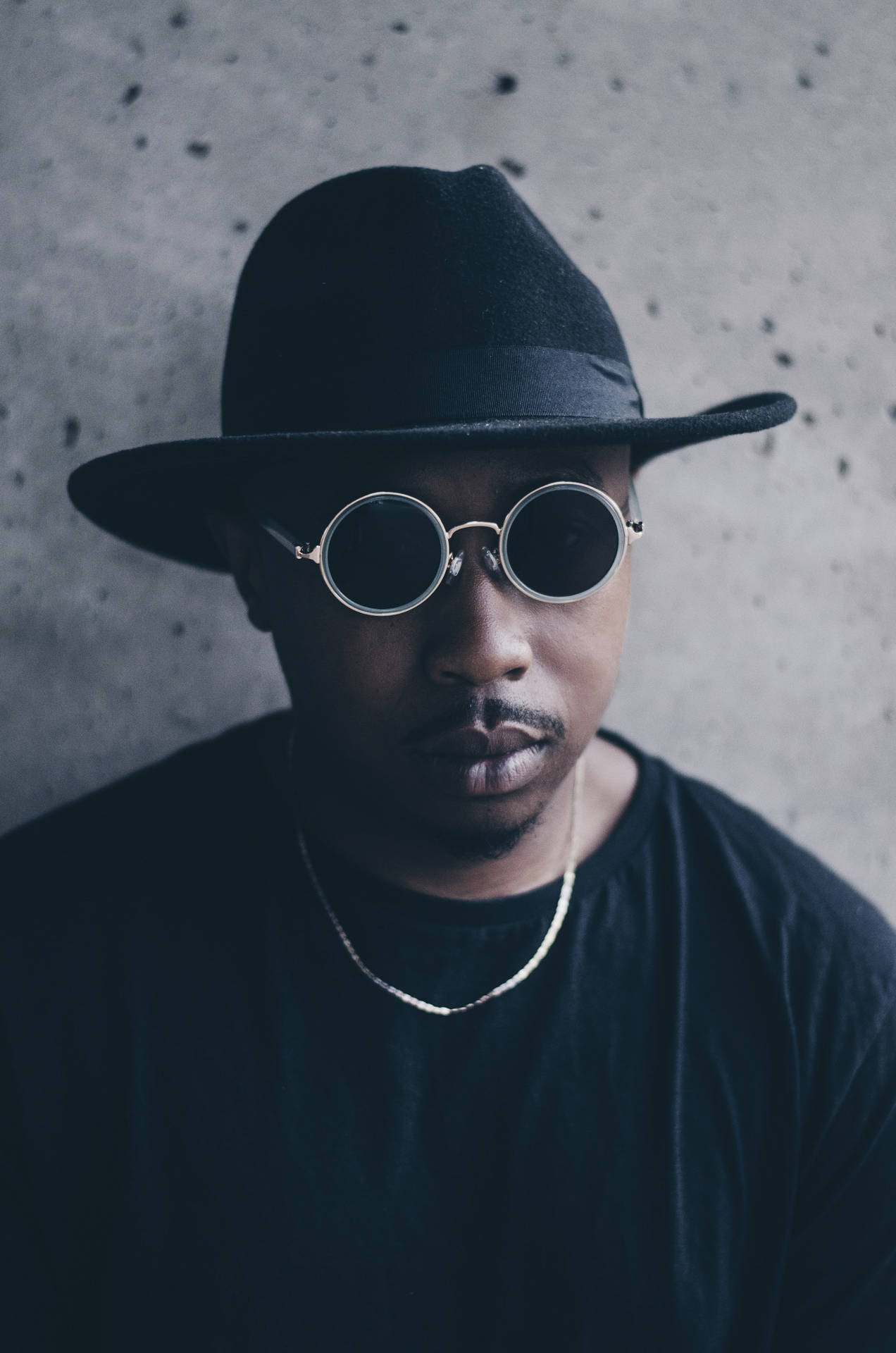 戴黑色軟呢帽和圓形太陽眼鏡的男人，在混凝土牆前。