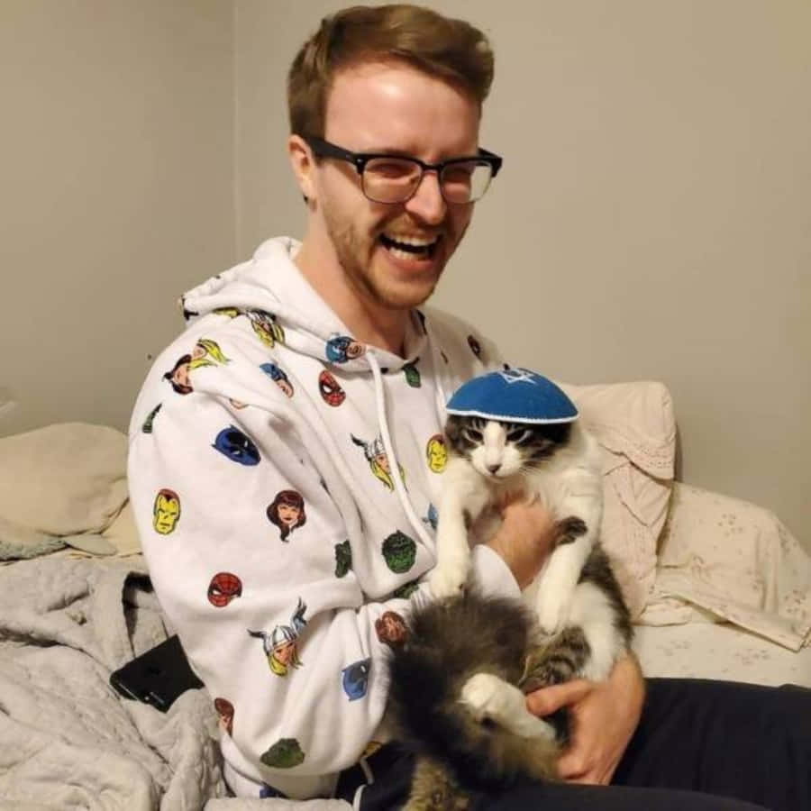 戴猶太帽的可愛貓咪主人 壁紙 — 一個快樂的男人穿著超級英雄連帽衫，抱著兩隻貓，其中一隻戴著藍色小圓帽，笑著。