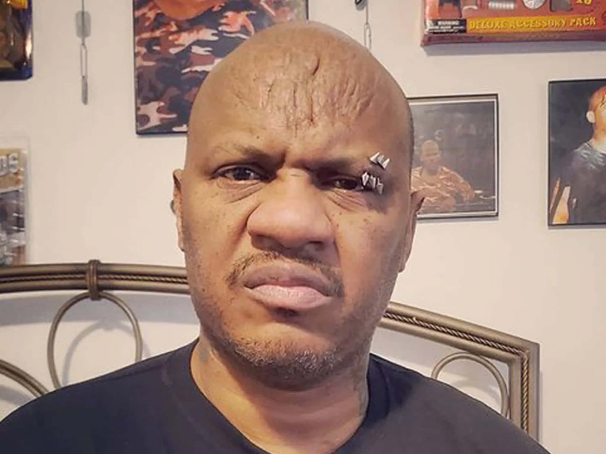 戴眉環的紐·傑克 壁紙 — 一個光頭、面部有穿孔的男人，在身後牆上掛有裱框海報的房間裡，強烈地凝視著相機。