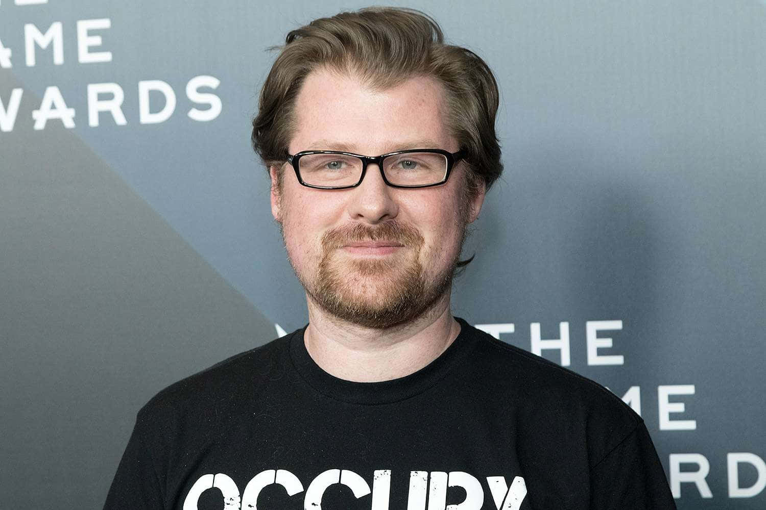 Justin Roiland 桌布 壁紙 — 戴眼鏡留鬍鬚的男人在Game Awards活動中穿著印有白色文字的黑色T恤對著鏡頭微笑。