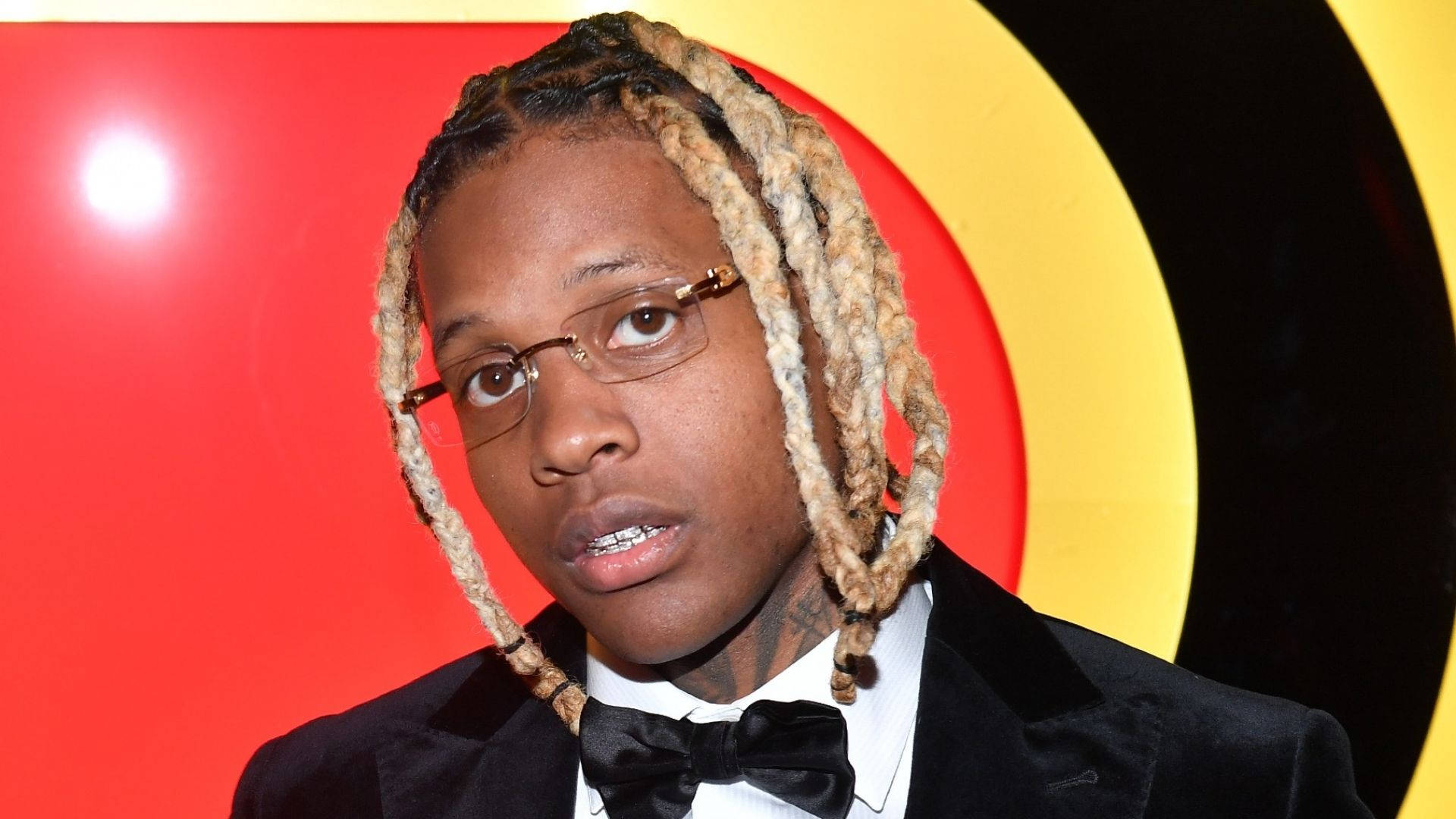 戴眼鏡的 Lil Durk 壁紙 — 一名留著辮子、戴眼鏡的男子，在搶眼的紅、黃、黑圓形背景前穿著黑色燕尾服。