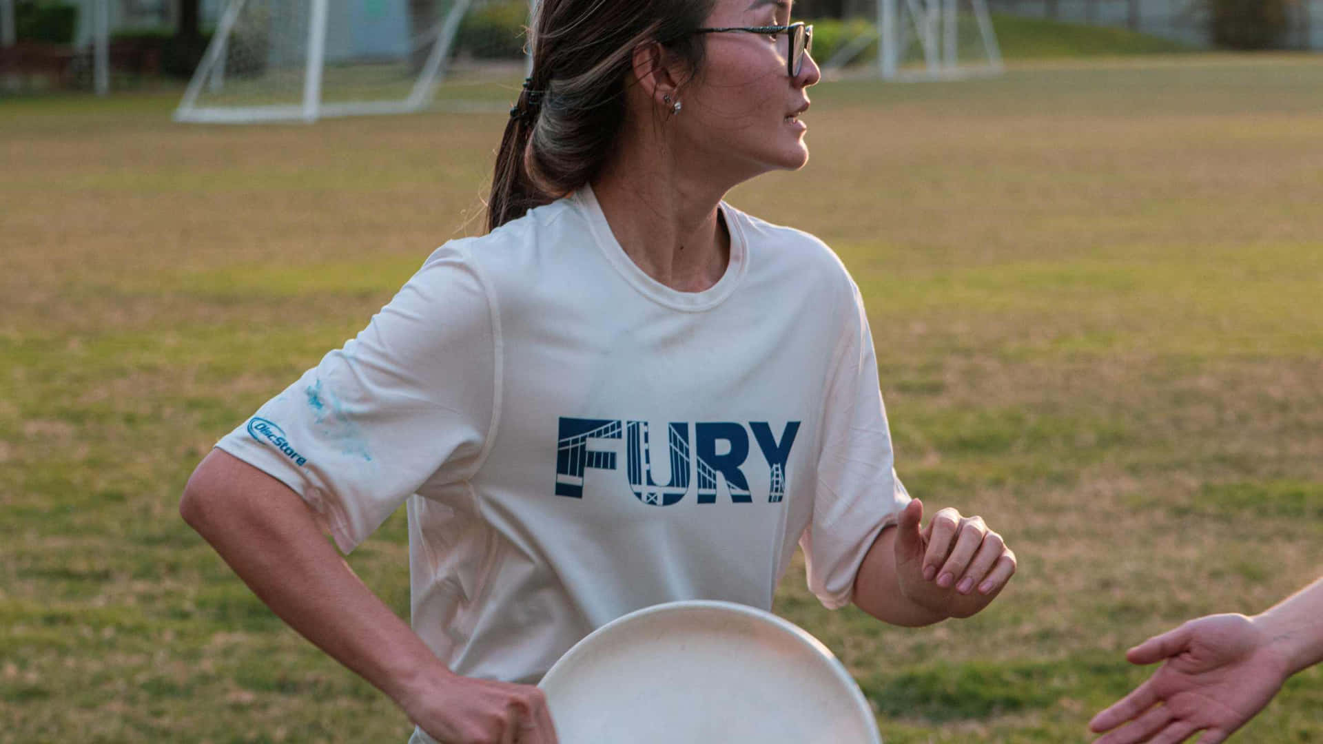 戴眼鏡的書呆女校園玩飛盤背景（1440p） 壁紙 — 一名身穿白色「FURY」T恤的女性在黃金時段的草地上拿著飛盤，背景有足球龍門。